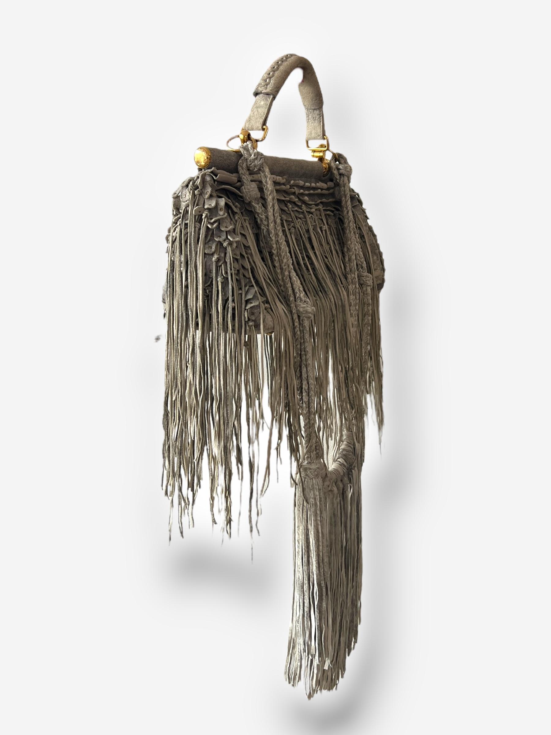 
Sac à franges en daim Roberto Cavalli 2011. Daim gris usé avec franges nouées et micro clous. Quincaillerie de couleur dorée. Un compartiment principal avec une poche latérale zippée. Deux poches extérieures zippées. Poignée supérieure avec