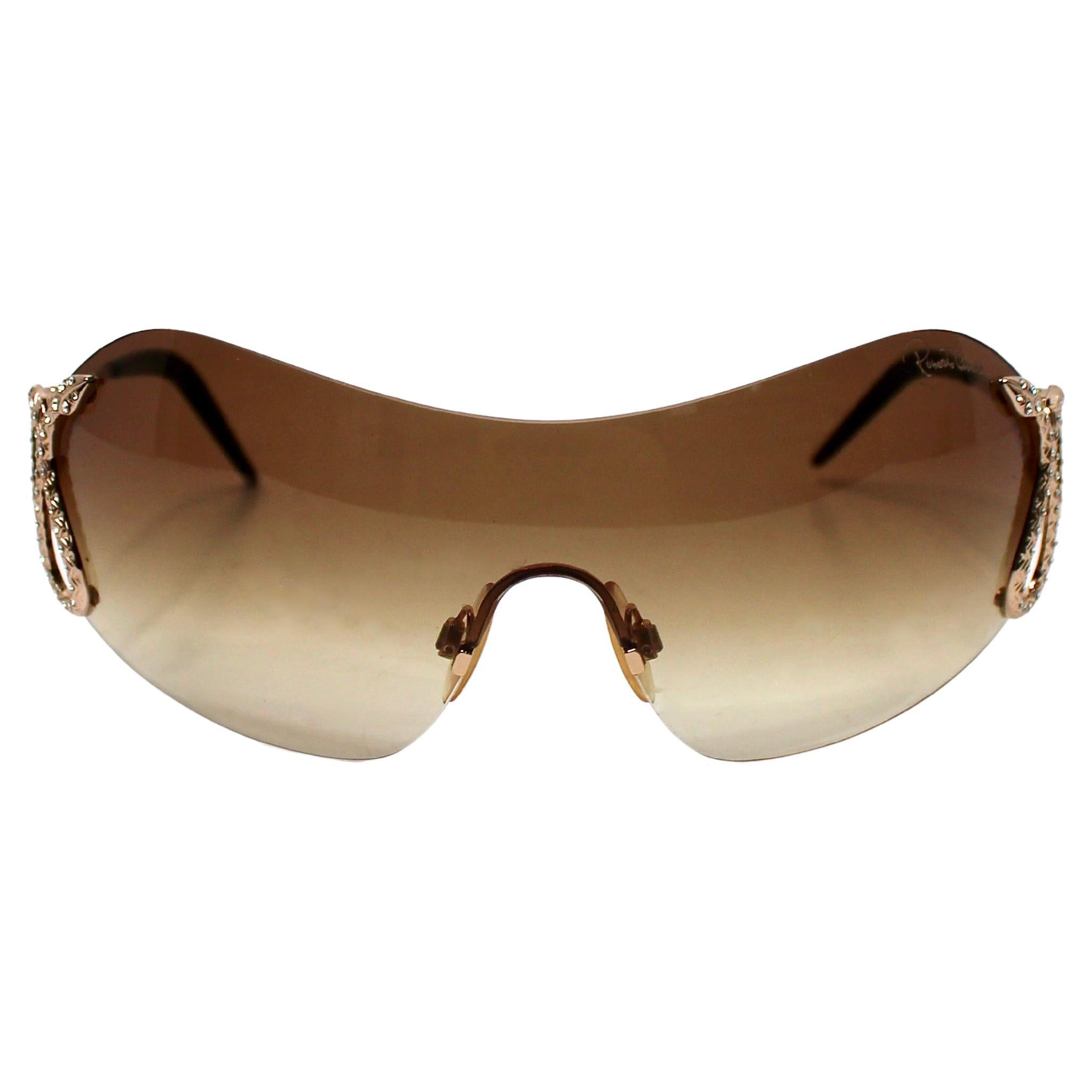 Questi occhiali da sole Roberto Cavalli con motivo a serpente con strass e scudo oversize, risalenti all'anno 2000, sono un pezzo audace e glamour che racchiude perfettamente l'energia vibrante dei primi anni 2000. La montatura a scudo oversize è