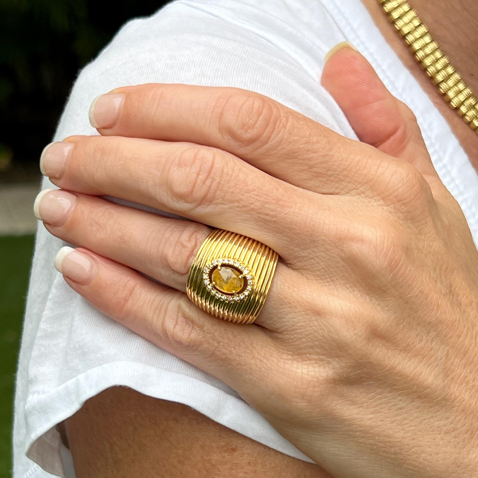 Yellow sapphire and diamond ring by Italian designer Roberto Coin. The ribbed wide band is fashioned in 18 karat yellow gold. The ring features an oval yellow sapphire gemstone weighing approximately 1.00 carat surrounded by 22 round brilliant cut