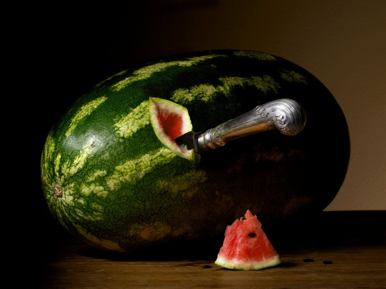 The First Cut by Robyn Stacey features a large watermelon sitting on a wooden table in this dramatically lit still-life. The handle of a silver knife protrudes out from a cut in the fruit. A chunk of watermelon sits next to the larger fruit below
