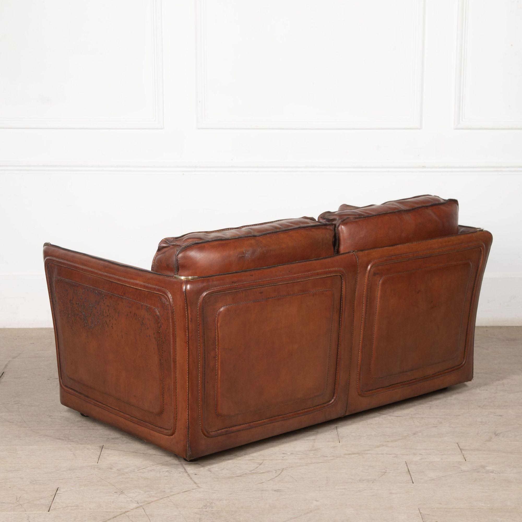 A stylish leather two-seater sofa is covered in sumptuous thick saddle leather with stitched edges and metal corner details.
The feather-filled cushions are extremely soft and comfortable.
circa 1950.