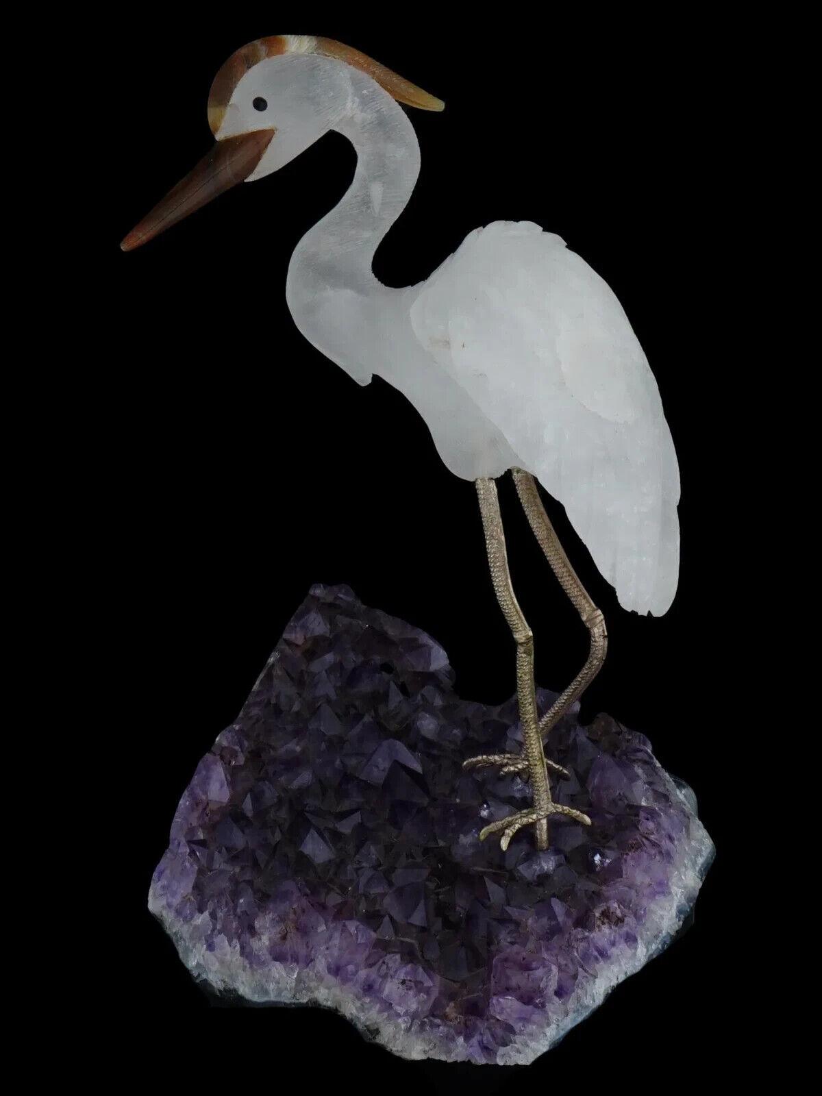 Sculpture d'oiseau héron en cristal de roche gravée à la main avec des motifs détaillés. Le bec et la crête de l'oiseau sont en agate. L'oiseau repose sur une grande géode d'améthyste naturelle. Les pattes de l'oiseau sont en métal. Figurines
