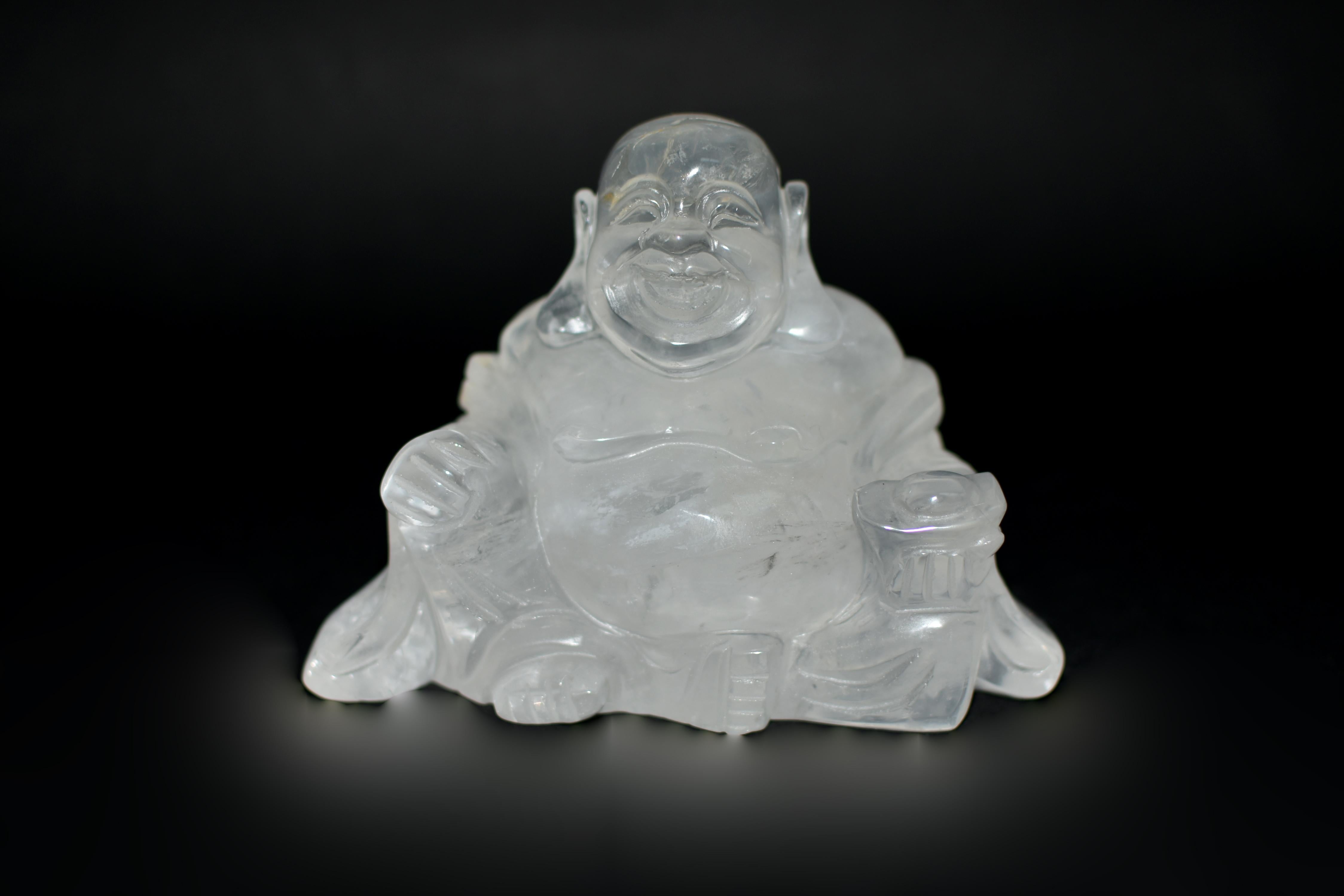 A precious quartz Happy Buddha, hand-carved from a solid block of all-natural fine grade rock crystal. Seated in serene repose, the Buddha's loose robe gracefully drapes around his upper body, cascading gently around his arms and legs. In his left