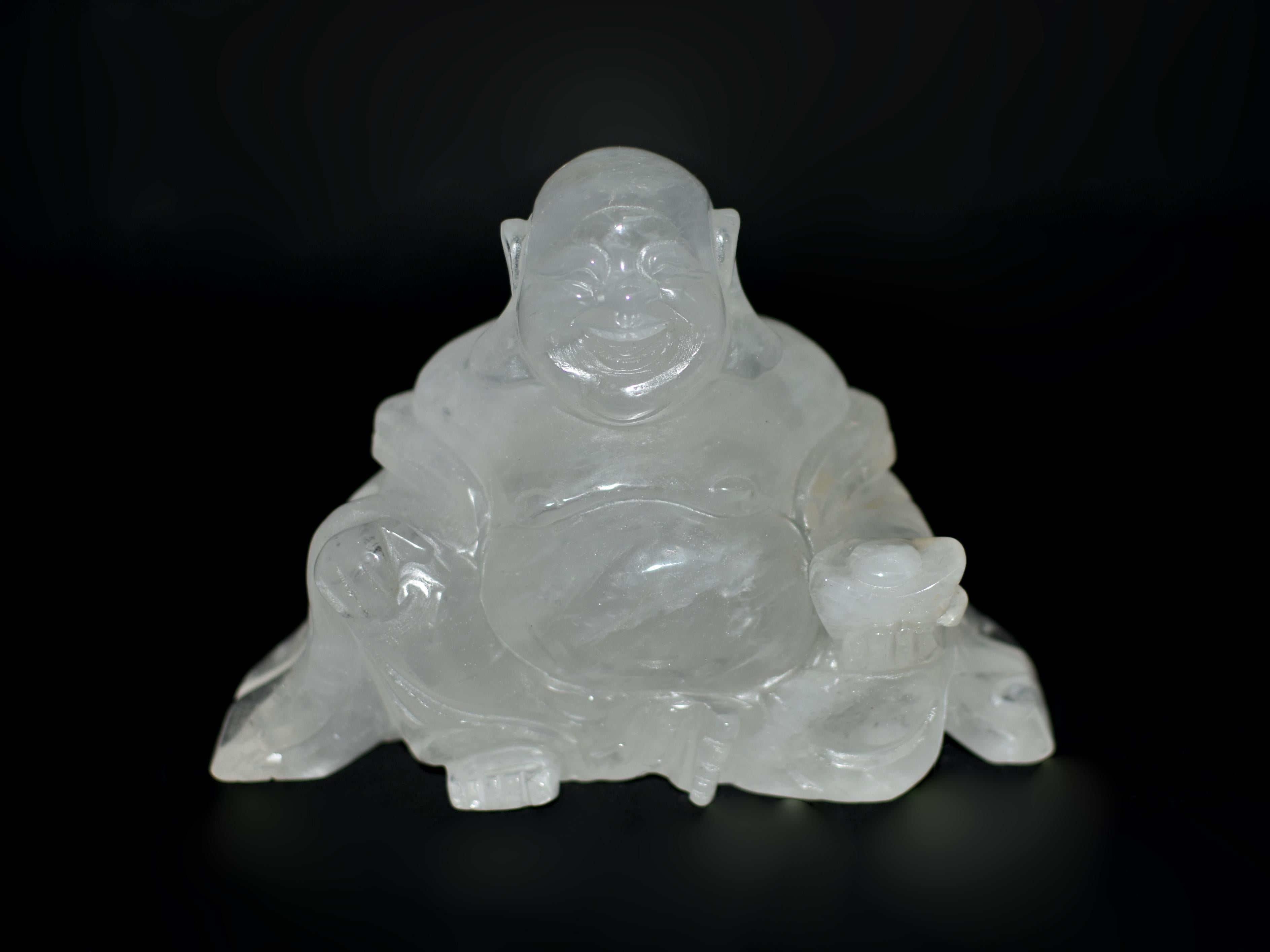 Ein kostbarer Quarz Happy Buddha, handgeschnitzt aus einem massiven Block aus natürlichem Bergkristall feinster Qualität. Das lockere Gewand des Buddha, der in heiterer Ruhe sitzt, legt sich anmutig um seinen Oberkörper und fällt sanft um seine Arme
