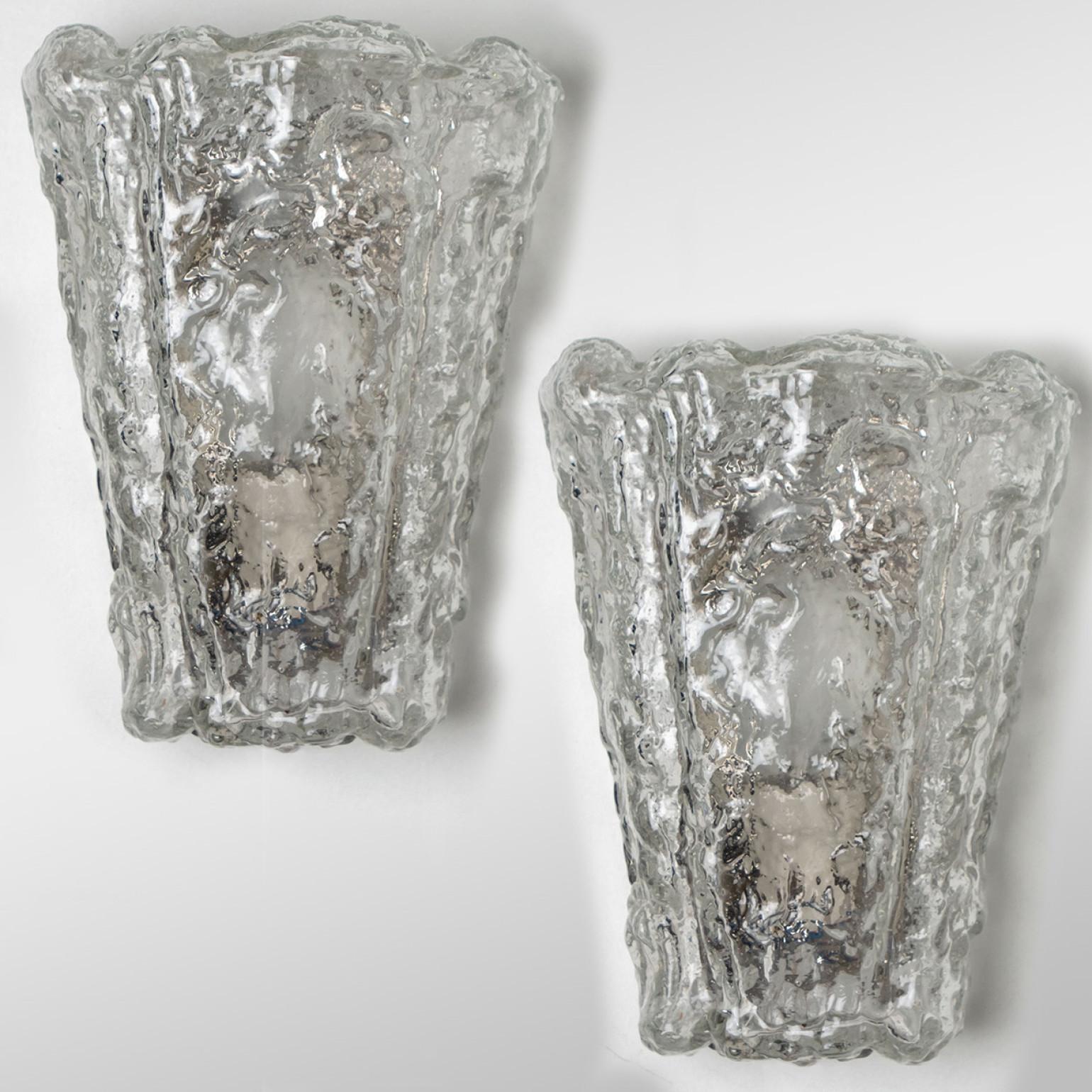 Pair of wall sconces by Hillebrand. Made in Germany around 1960.
The glass shade looks like a clear piece of rock. The glass refracts the light beautifully, filling a room with a soft glow.
The wall light features a black back plate and chrome