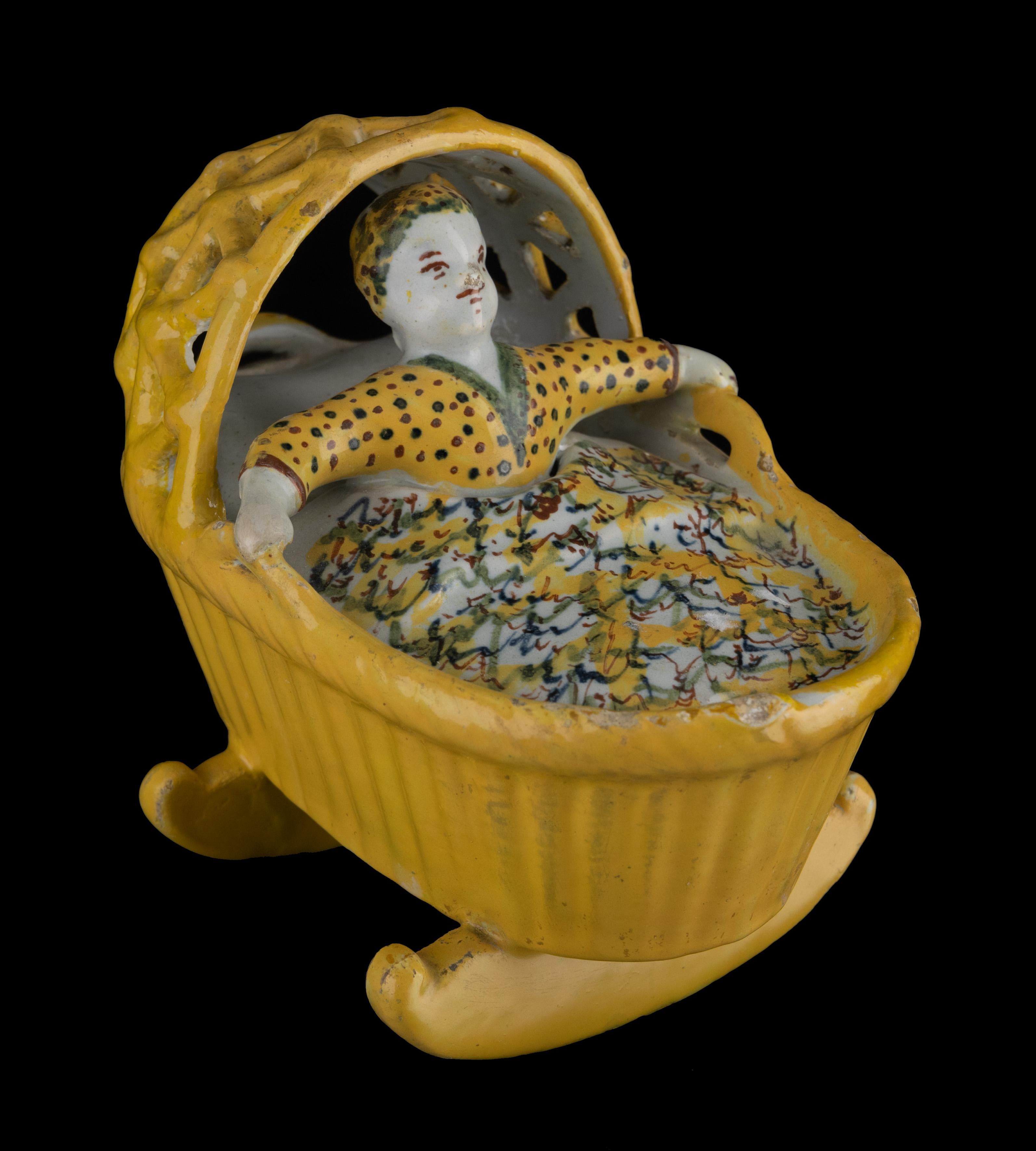 Rocking cradle with child. Delft, 1750-1780

Polychrome figure of a child in a rocking cradle with open hood. The child is dressed in a yellow nightgown and sleeping cap with reddish-brown and green dots. It sits upright under a marbled blanket