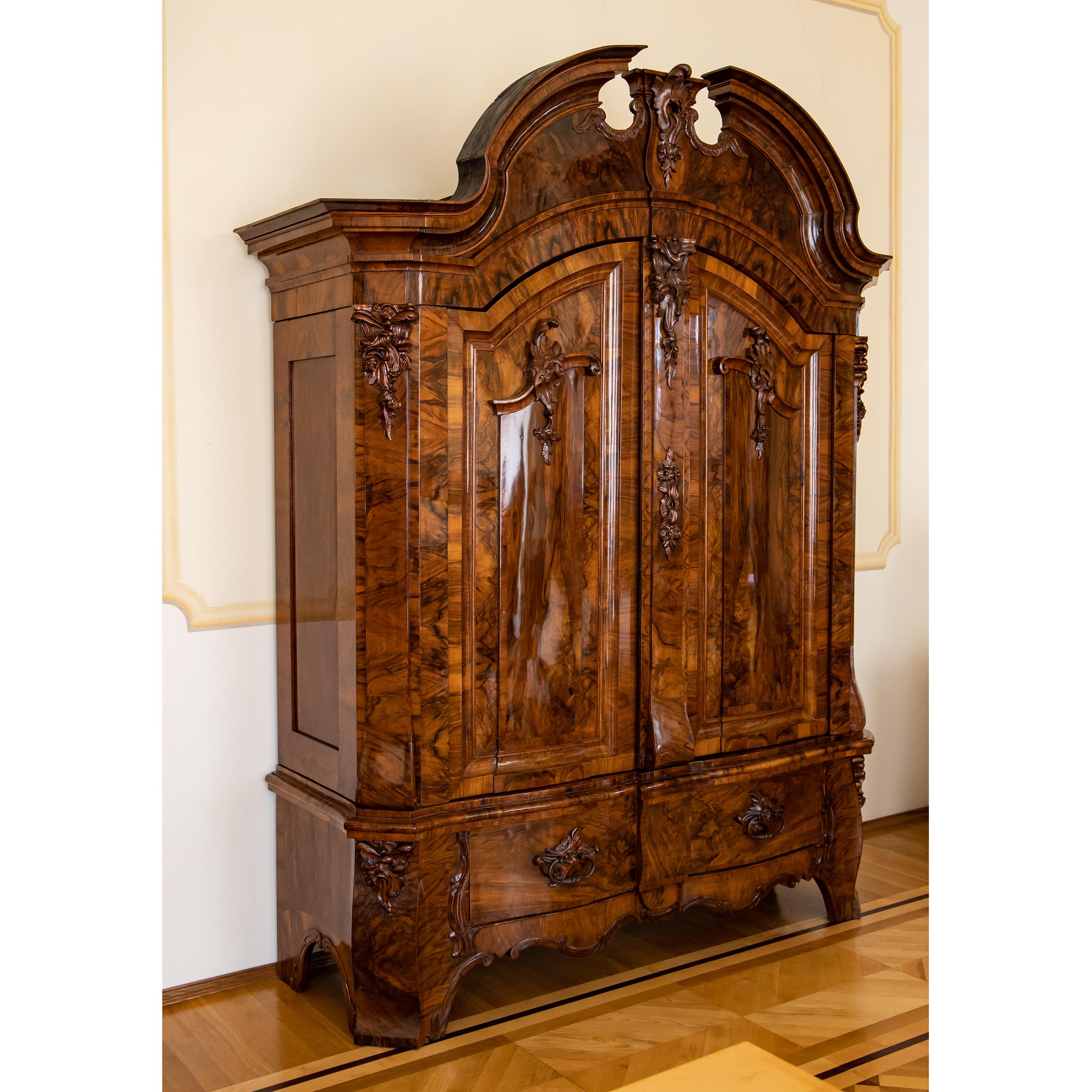 Large courtly hall cupboard with tall swan's neck pediment, bevelled corners and strongly pronounced plinth zone with two drawers and a multiple curved frame. The front is slightly wavy, the fillings of the doors are profiled and close arcade-like.