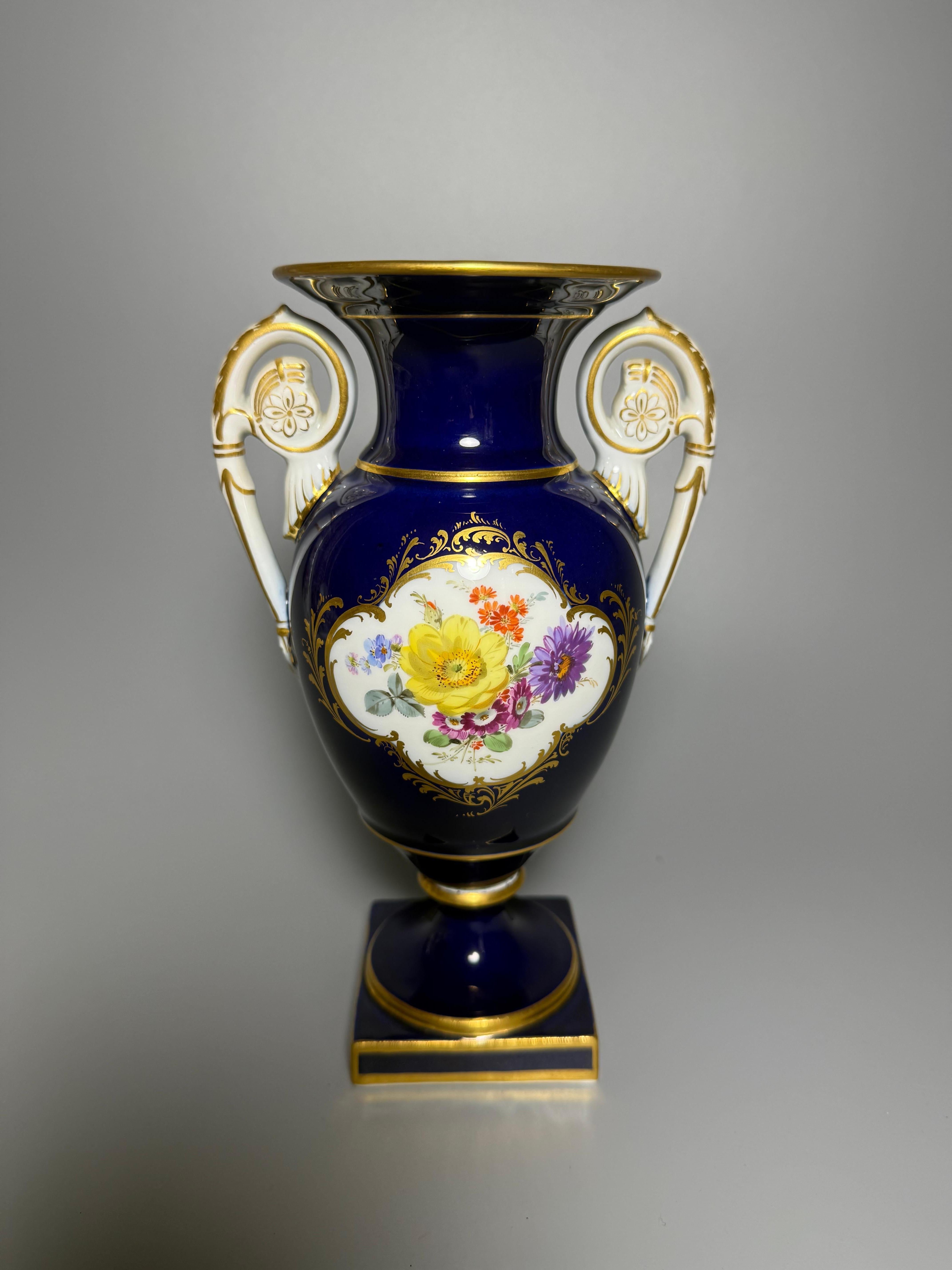 Meißener Amphora Vase
Diese Porzellanvase hat einen kobaltblauen Hintergrund mit aufwändigem Golddekor und polychromer Blumenmalerei. 
Sie steht auf einem quadratischen Sockel mit rundem, ansteigendem Fuß, der einen balusterförmigen Korpus trägt.