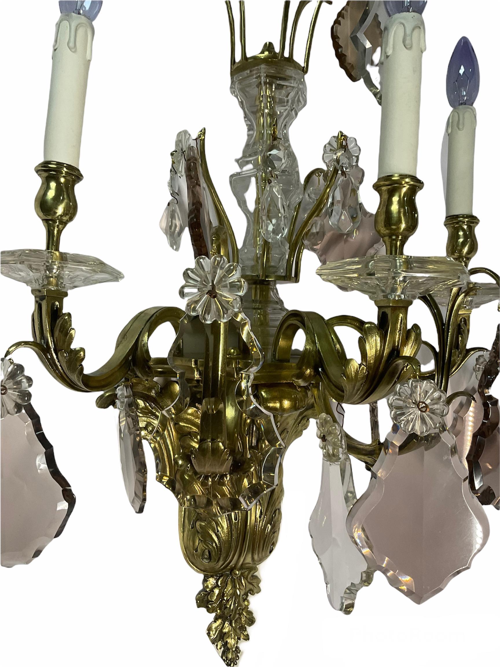 This is a Rococo Louis XV style pair of gilt bronze wall sconces. Their main body is made of three short bronze arms at the top embellished with clear pressed crystal rosettes and pendalogues over a cut crystal urn shaped tower that cover a gilt