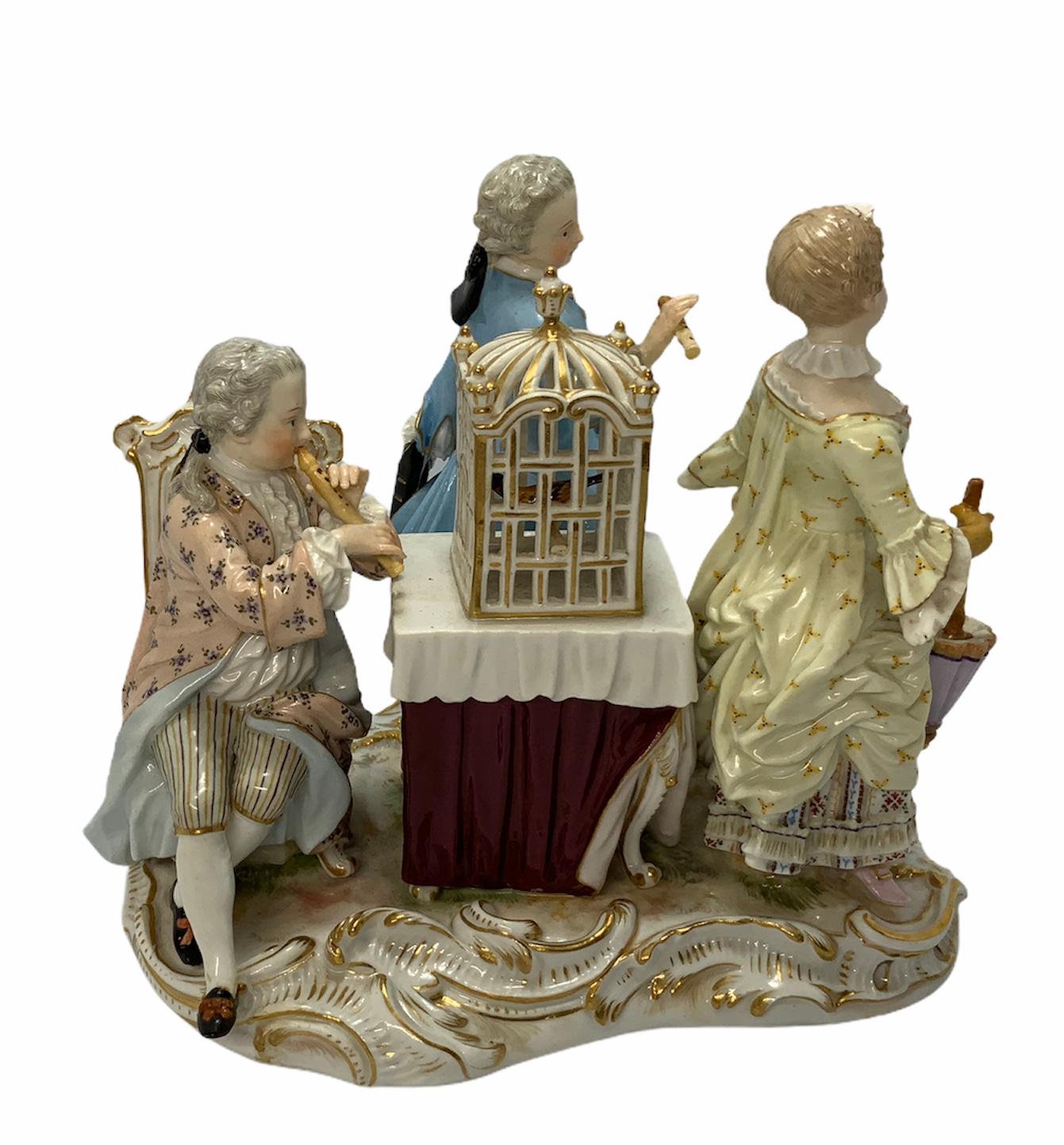 Meissen Porcelain group of figurines depicts a trio of children attire with beautiful 18th clothes around a birdcage over a table. There are two boys with flutes, one of them is sitting and playing it and the other is standing and holding it. There