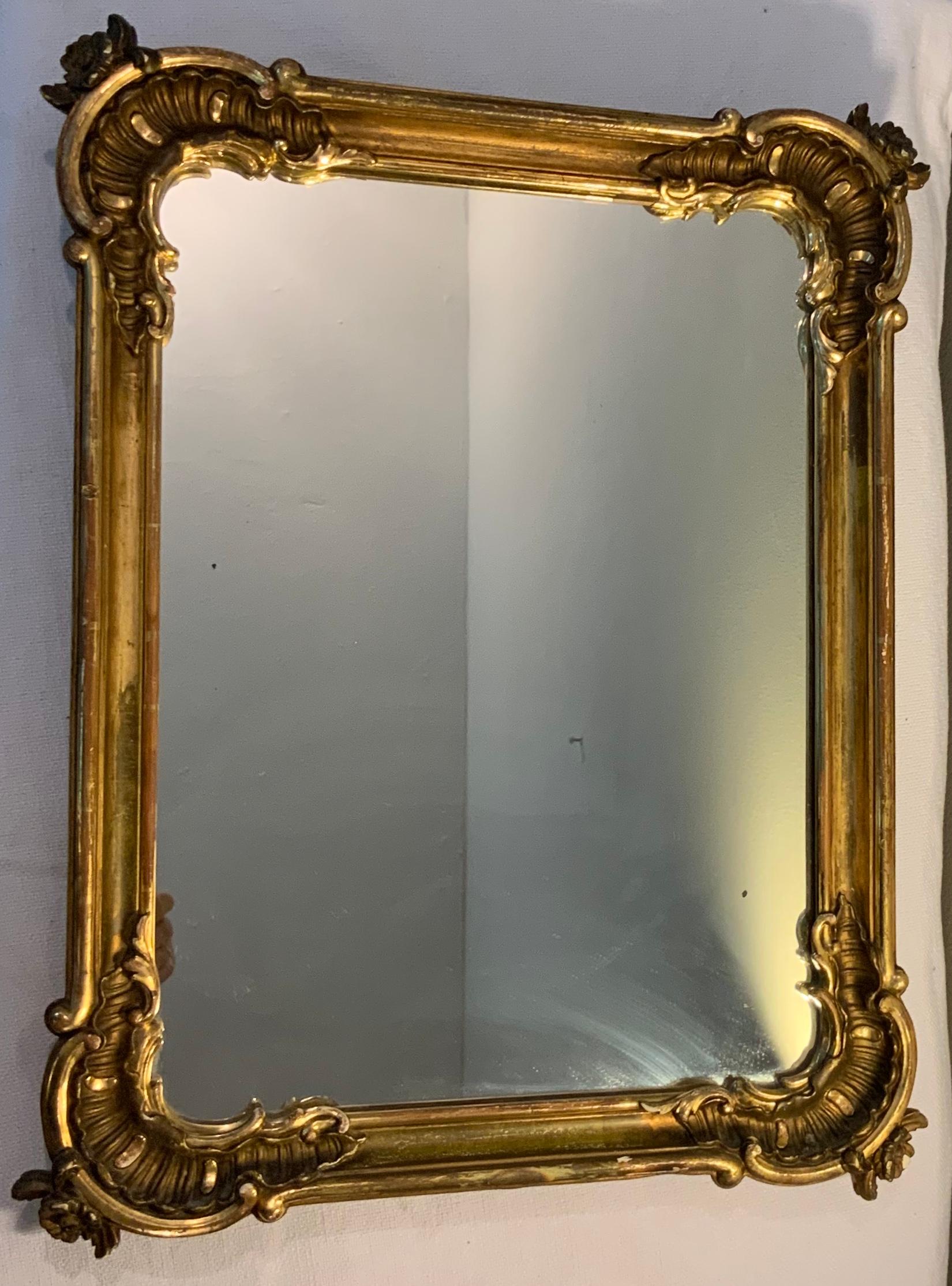 This a heavy rectangular gilt plaster-wood framed mirror adorned with large carved rocailles in the joints. Delicate scrolls delineate the borders and a rosette embellishes the top of each rocaille. There is a metal triangle hook in the upper back