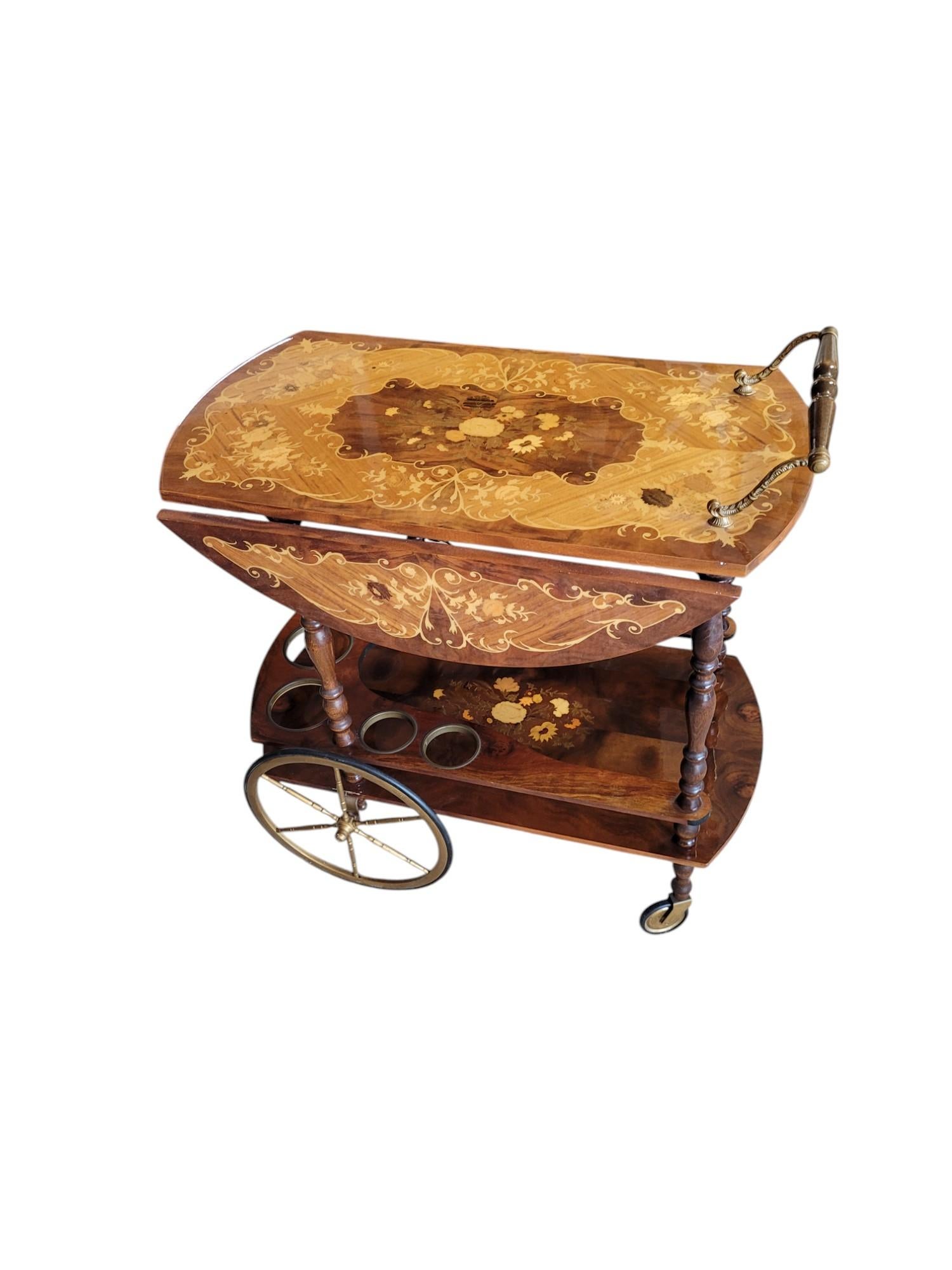 Elegante e funzionale, questo carrello da bar vintage presenta un piano con ante a ribalta, un raffinato intarsio floreale e una finitura lucida e brillante. Le gambe tornite, i portabottiglie bordati in ottone e le grandi ruote a raggi aggiungono