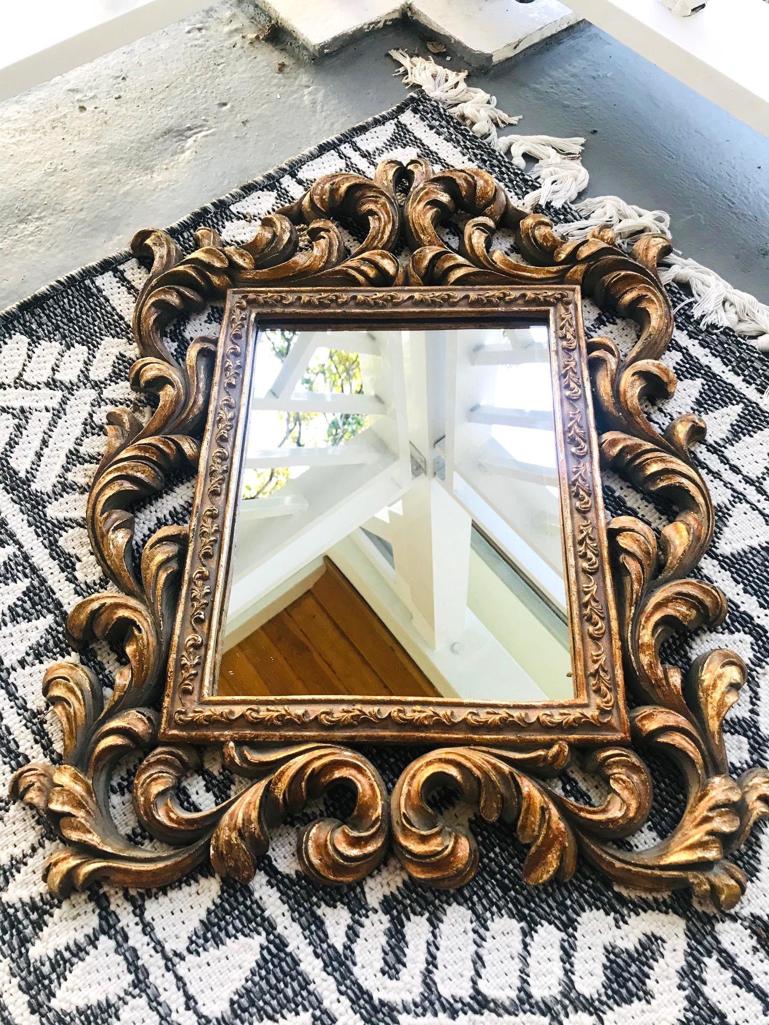 Hollywood Regency Baroque style mirror features a solid wood frame with hand carved designs and antique gold leaf finish. The ornamental frame features stylized scrolls of foliage and Prince of Wales plumes. The rectangular inner frame has a series