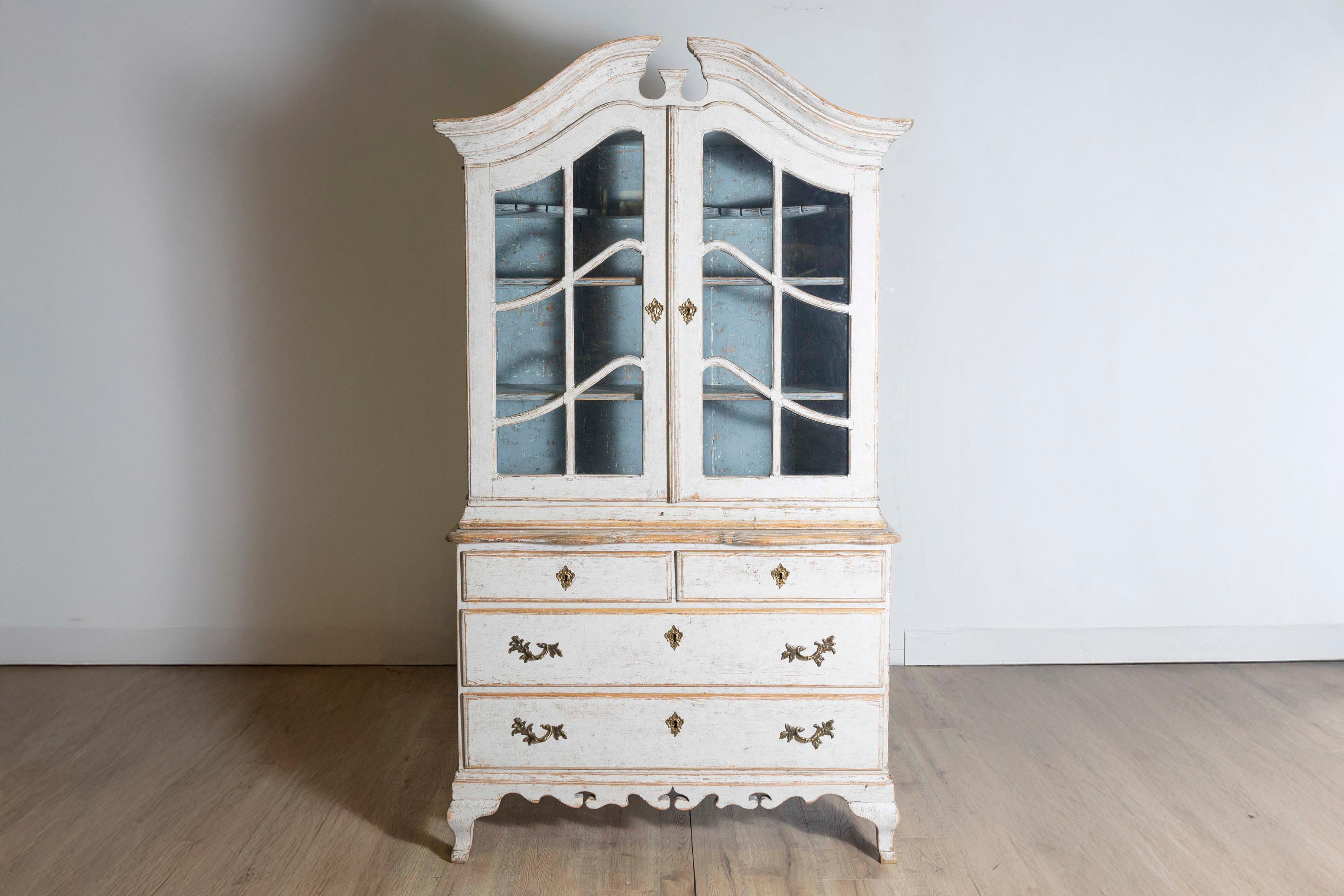 Ein schwedischer Vitrinenschrank im Stil des Rokoko, hellgrau lackiert, um 1850, mit Haubengiebel, Glastüren, vier Schubladen, geschnitzter Schürze und geschwungenen Füßen. Verströmen Sie Eleganz mit diesem schwedischen Vitrinenschrank im Rokokostil