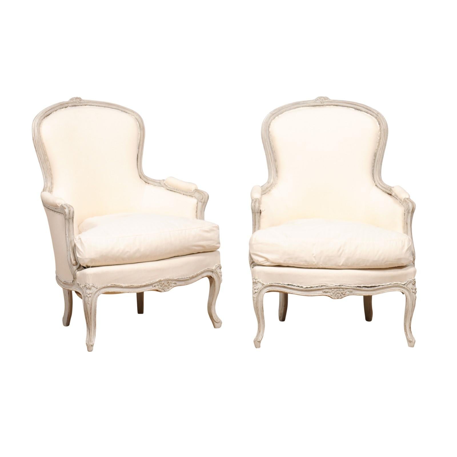 A pair of Swedish Rococo style classic light grey painted bergères chairs from circa 1890 with carved floral décor, cabriole legs and upholstery. Immerse yourself in the poetic allure of this pair of Swedish Rococo Style bergères, circa 1890,