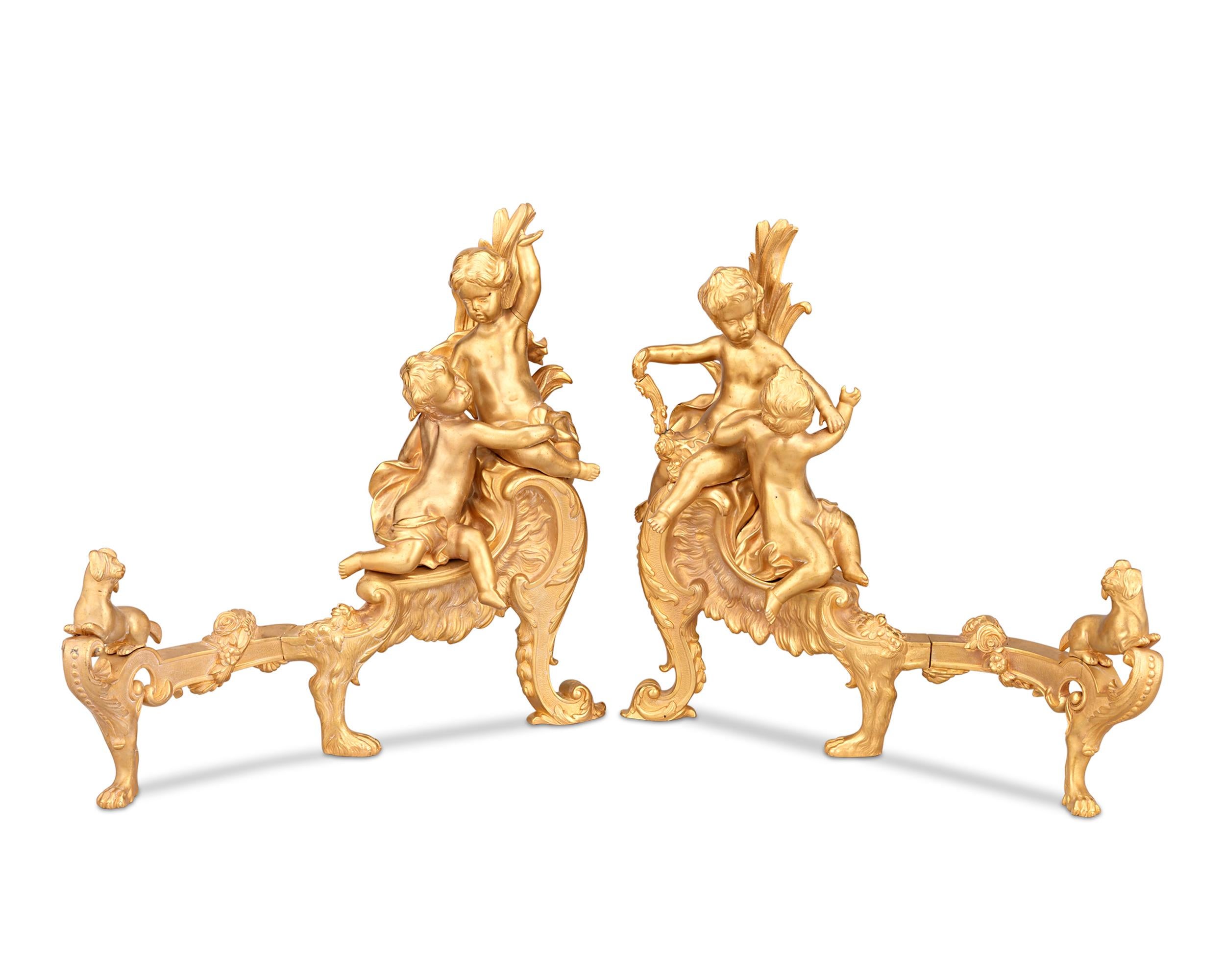 This charming pair of Rococo-style doré bronze French chenets features charming cherubs seated out their ornate plateau. Canine feet hold the chenets aloft, while two dog figures adorn the ends. Chenets, also known as andirons, were once staple of