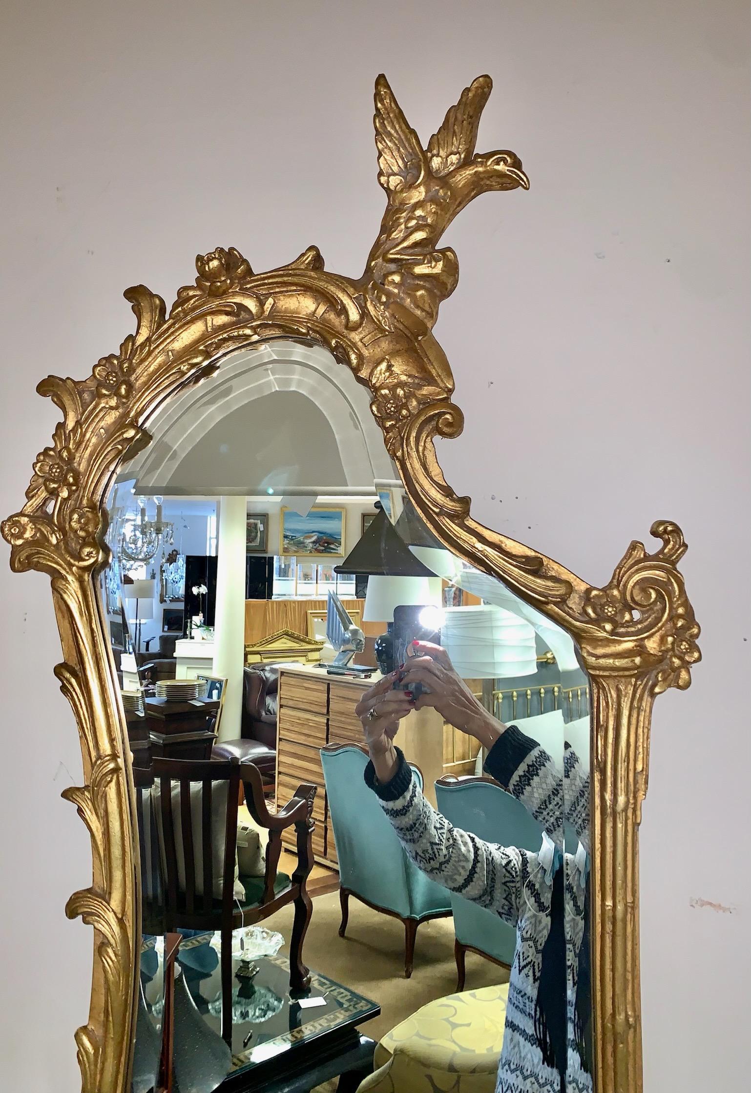 Magnificent pair of ornate giltwood shaped mirrors feature a surmounted carved phoenix bird with floral and foliage decoration enclosing a beveled mirror. Each has a projected two arm candle sconces. By Friedman Brothers.