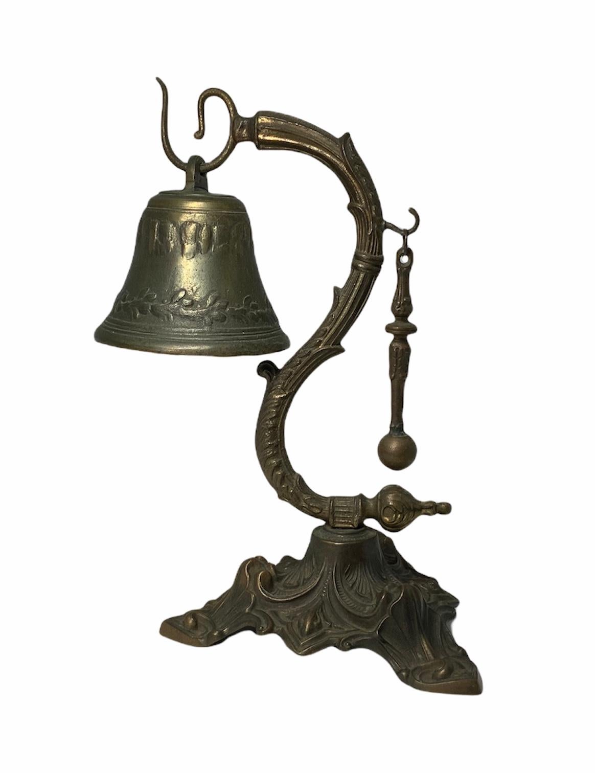 This a bronze color heavy white metal call bell with a stand and a small bell stick. The bell is adorned with some repousse bay leaves branches. It hangs from a hook in a stand made of a serpentine shaped acanthus leaves intertwined branches. This