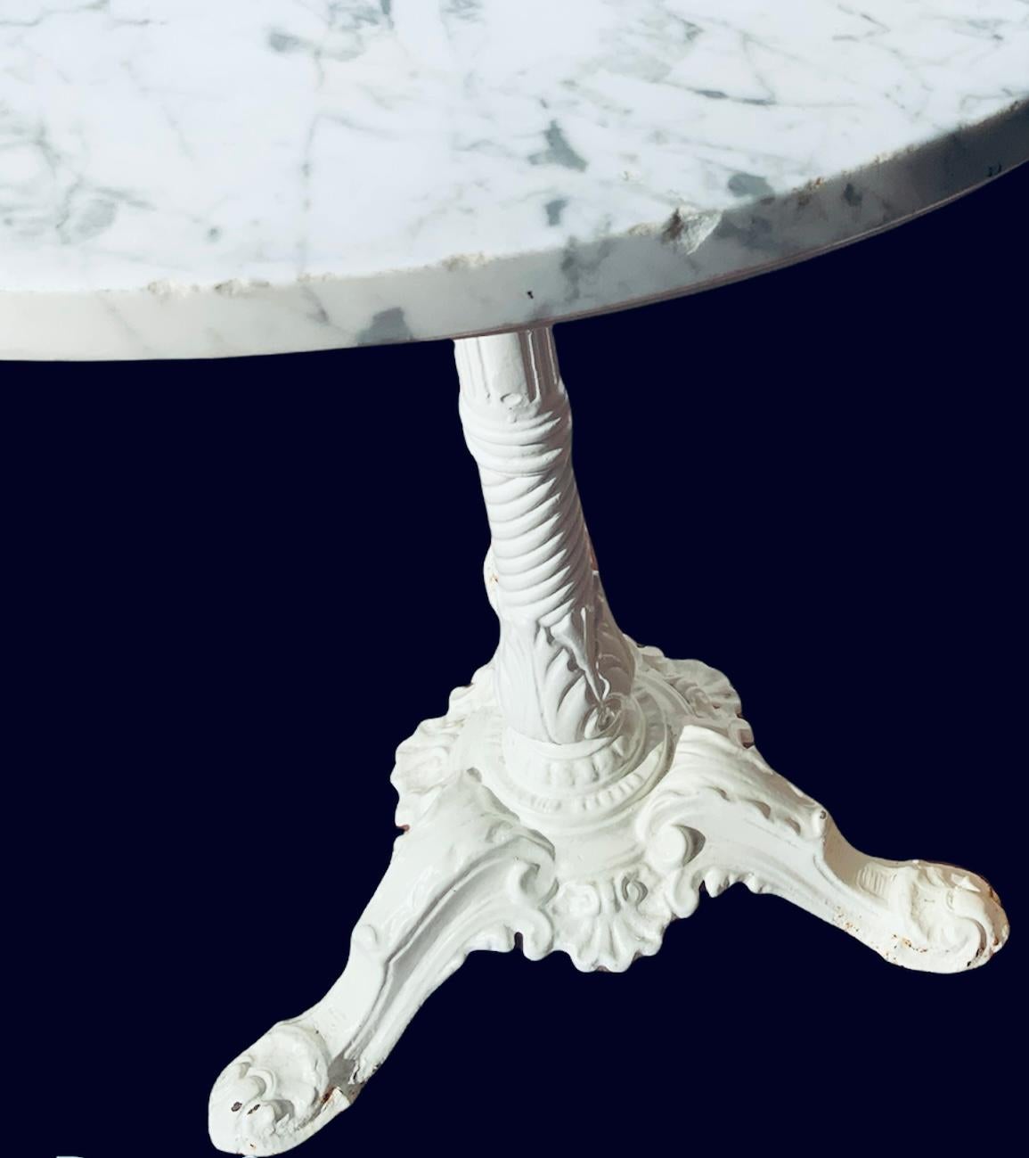 This a round white marble top cast iron bistro table. The marble top has grey veins and it is over a round cast iron base that is supported by a pedestal. This one consists of a vertical fluted lines at the upper top of the column and ending with a