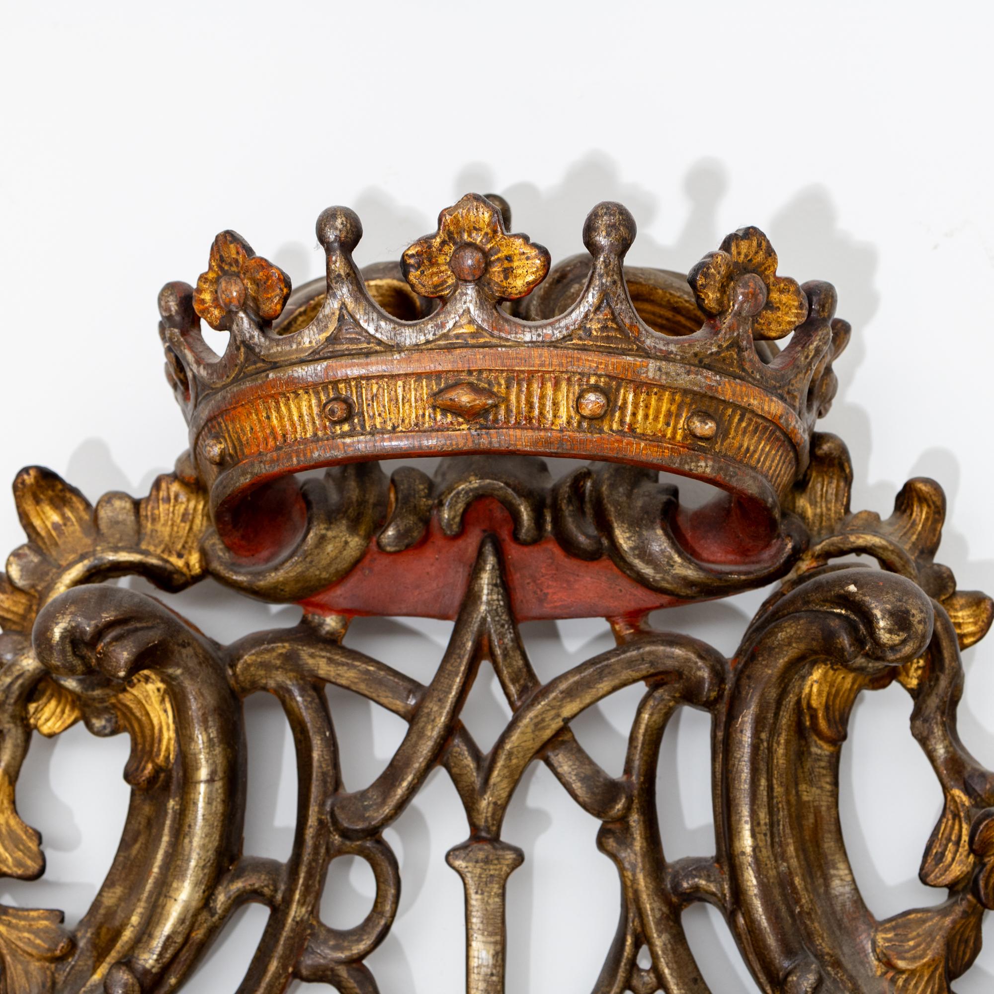 Rococo wall mirror in richly carved, silver and gold patinated wooden frame with rocailles and shell motifs. The frame is crowned by the three-dimensional crown protruding into the room above the monogram of the Virgin Mary.