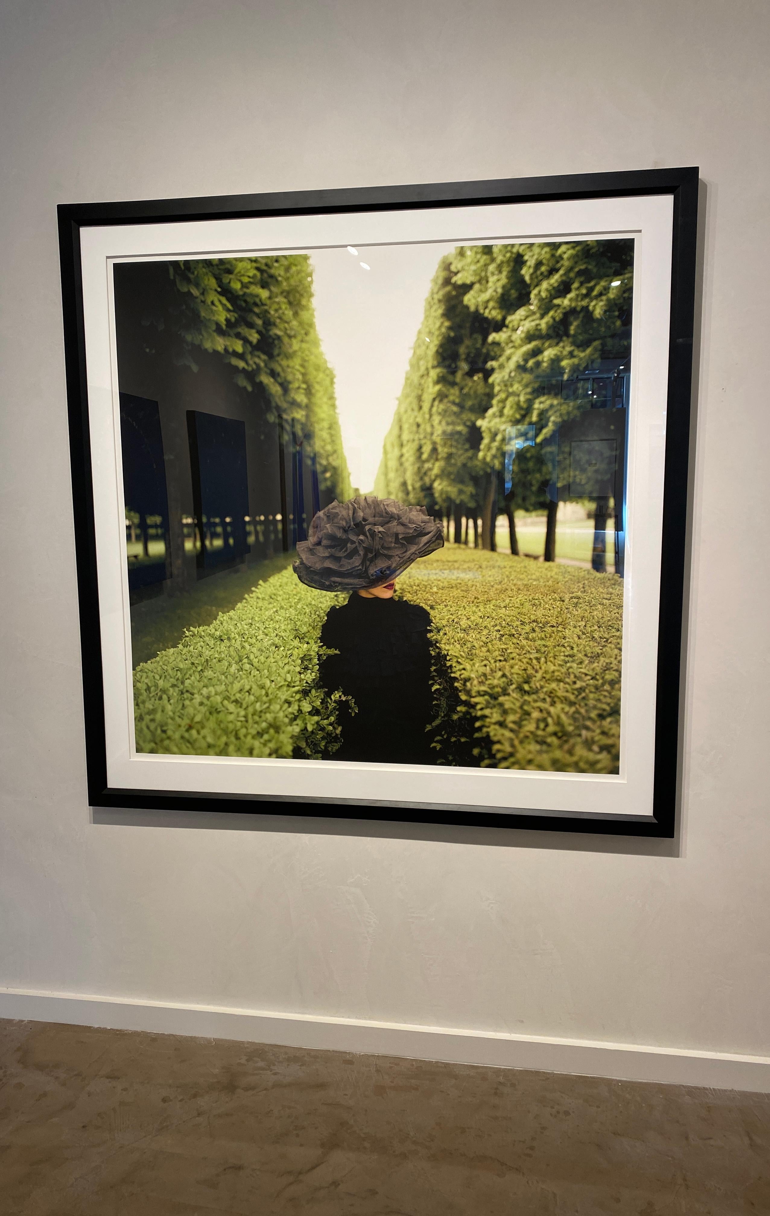 Color photograph of a woman in a wonderfully large and sculptural hat between lush green hedges in Paris.
Image size: 50 x 50 inches 
Framed size: 60 x 60 inches
Each image is part of an edition of 25. Price increases as the edition sells out. His