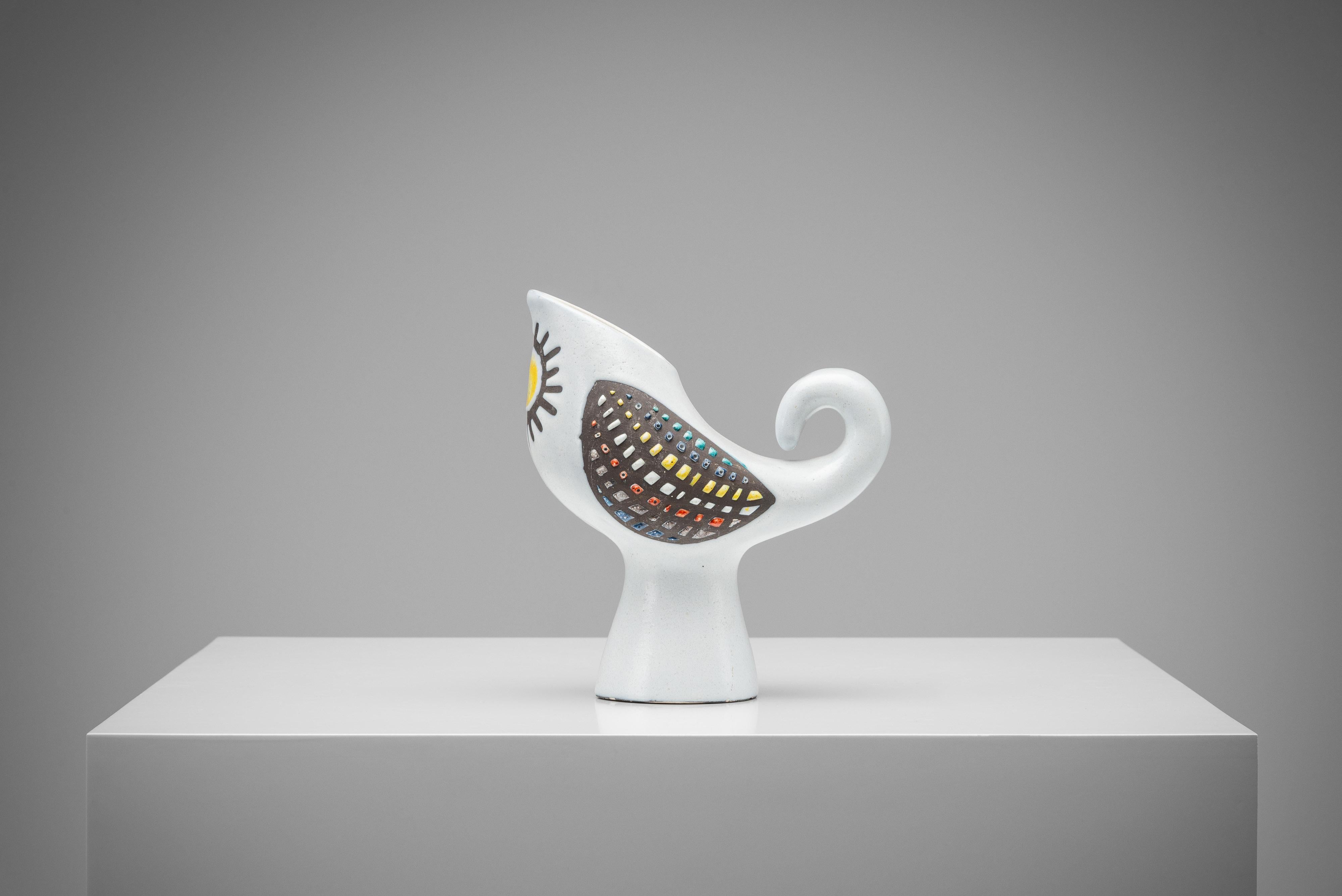 This ceramic pitcher is a special creation by Roger Capron and made in France in 1960. It's shaped like an abstract bird or rooster and was made in the 1960s. It has a surprising design with a soft white glaze and colourful, tile-like wings. This