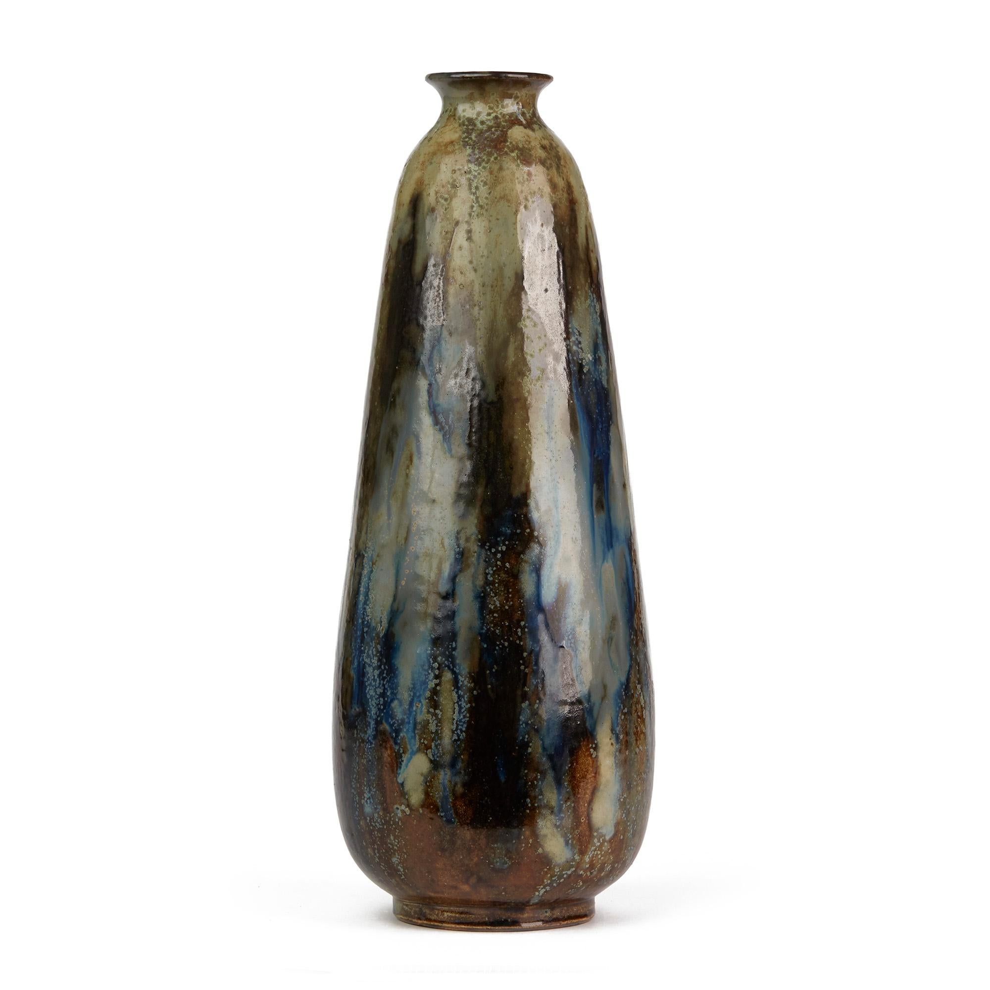 A stunning and exceptionally glazed Belgian art pottery stoneware vase by Roger Guérin and dating from circa 1930. This tall bottle shaped vase stands on a narrow rounded base and is decorated with colored streaked glazes in tones of brown, blue and