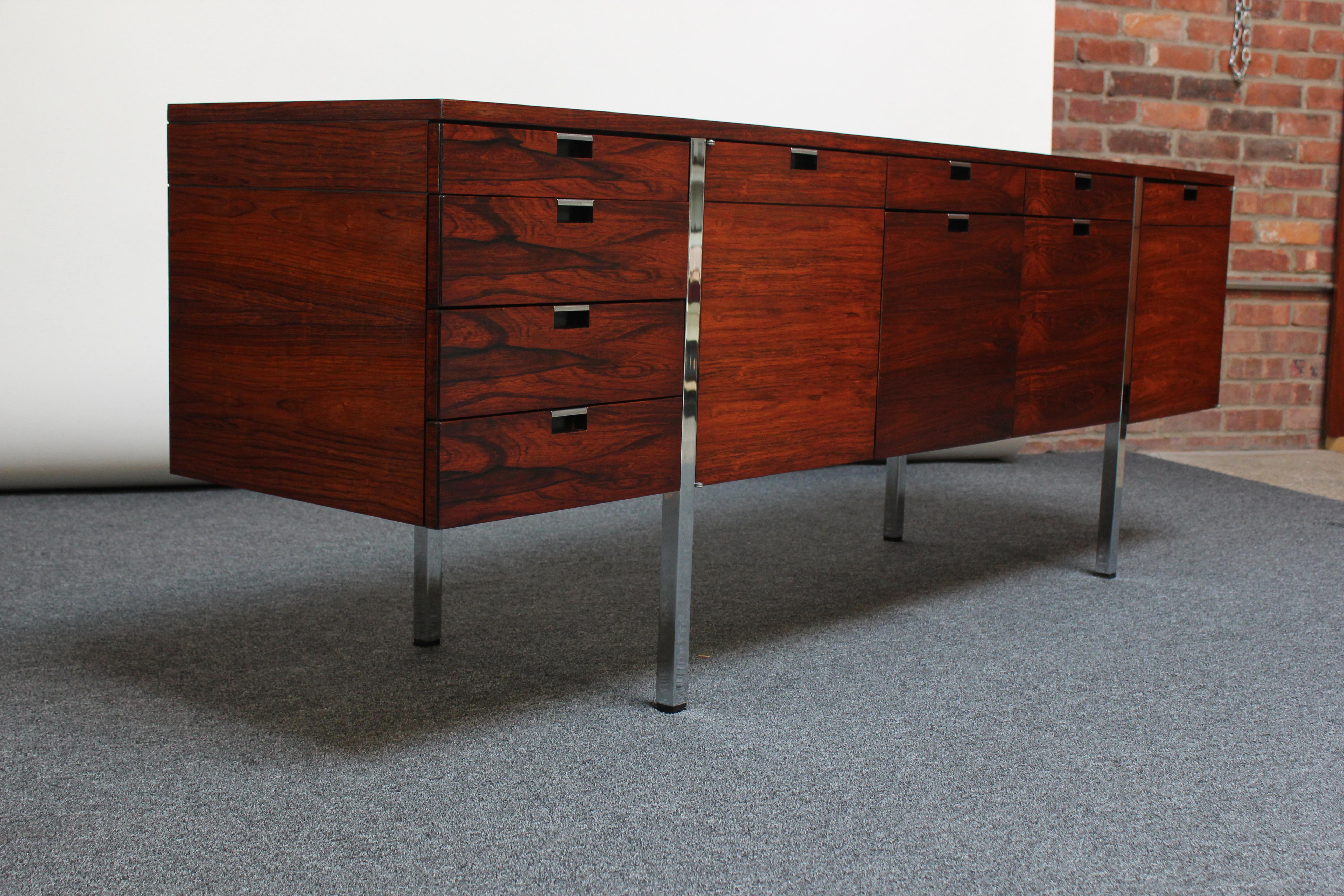 Handsome credenza designed in the 1970s by Roger Sprunger for Dunbar composed of exquisite rosewood veneer and chrome inset pulls and legs. 
Ample storage in a variety of configurations: far left side features one shallow drawer atop three more