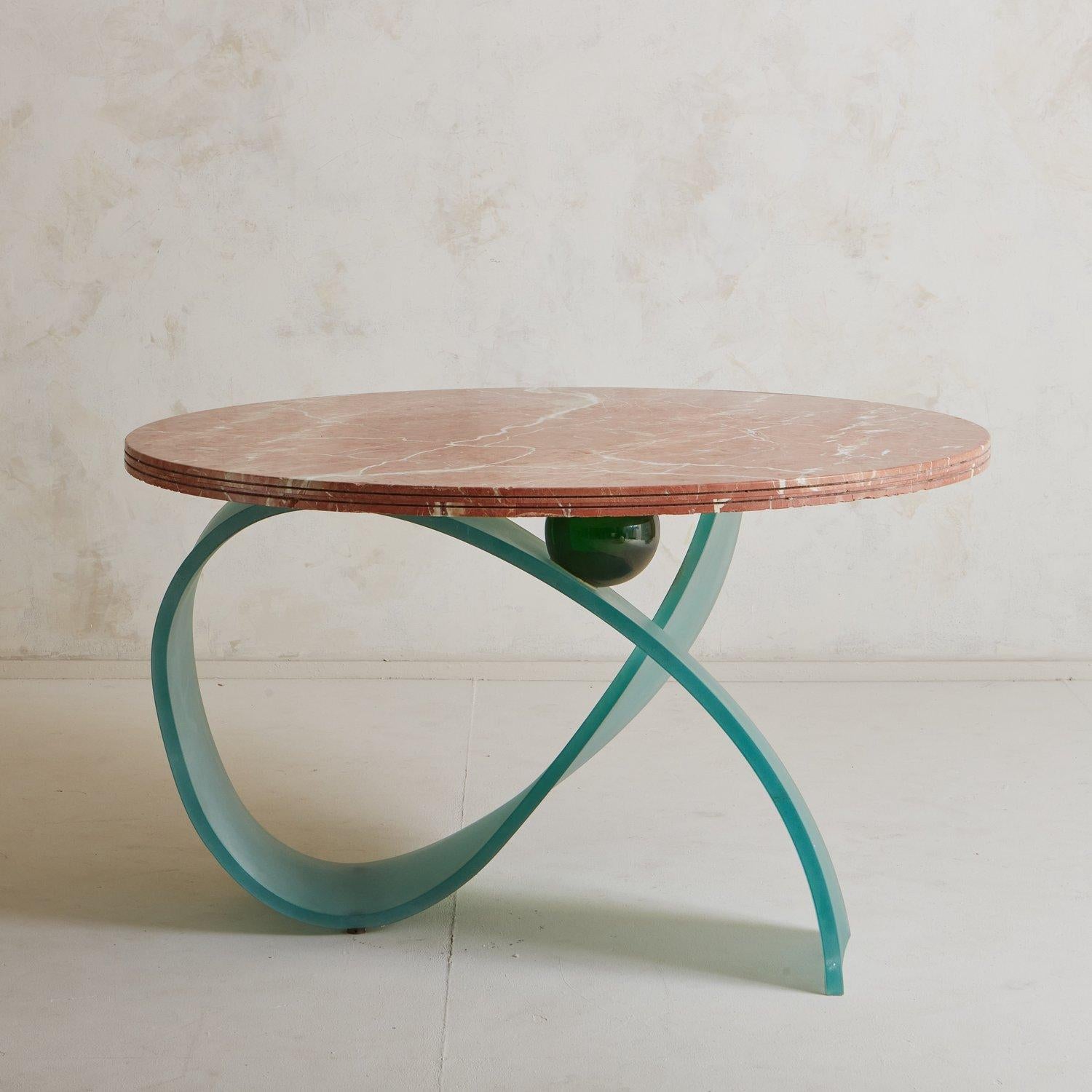 A vintage dining table with a round table top constructed with three layered blocks of Spanish Rojo Coralito marble featuring gorgeous cream and gray veining. This unique table has a sculptural blue frosted glass base with a green sphere detail.