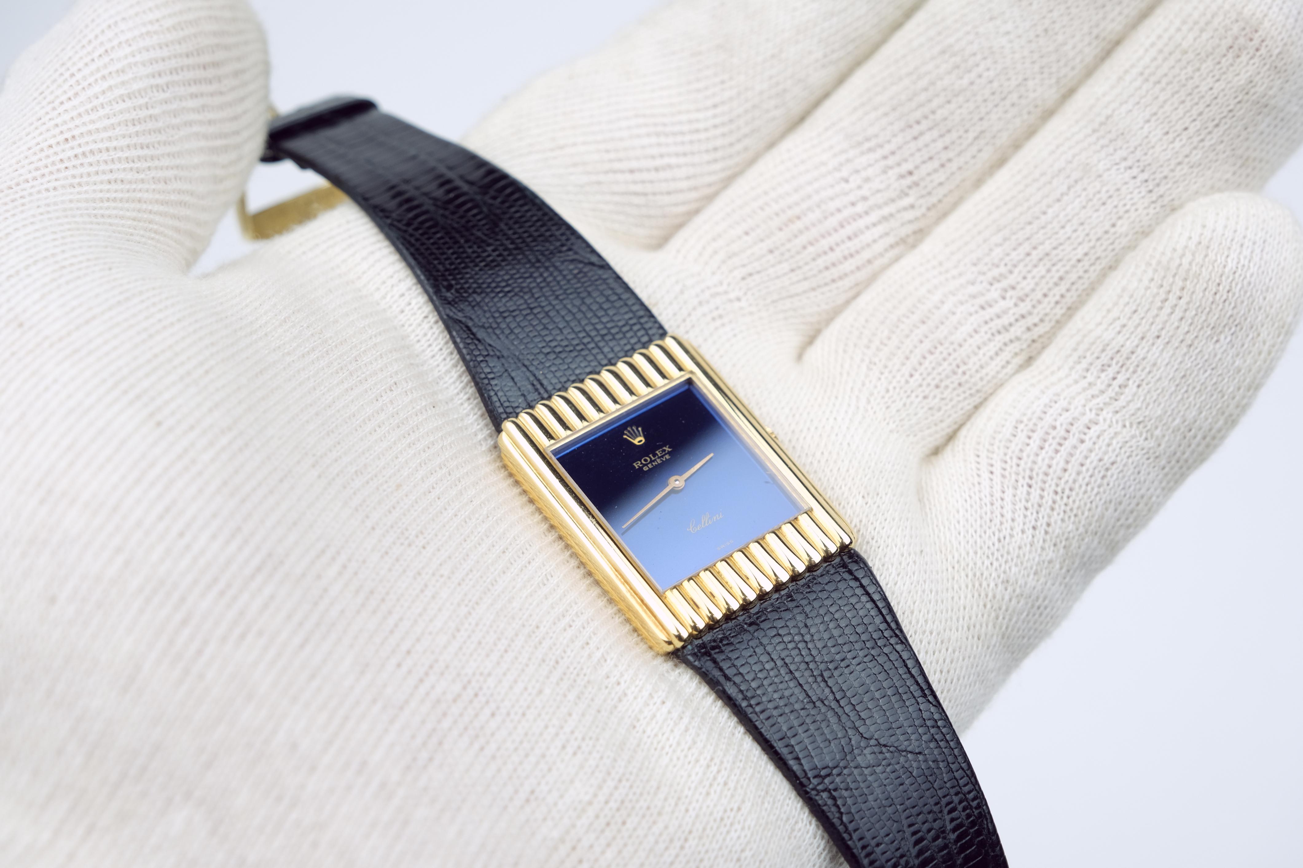 A Rolex Cellini 18K Gold Stem Wind Wristwatch. A luxury timepiece with a tank-style 18K yellow gold bezel that frames a navy blue dial with Rolex hallmark crown and gold-tone baton-styke hands.

This luxury watch also features an 18K yellow gold