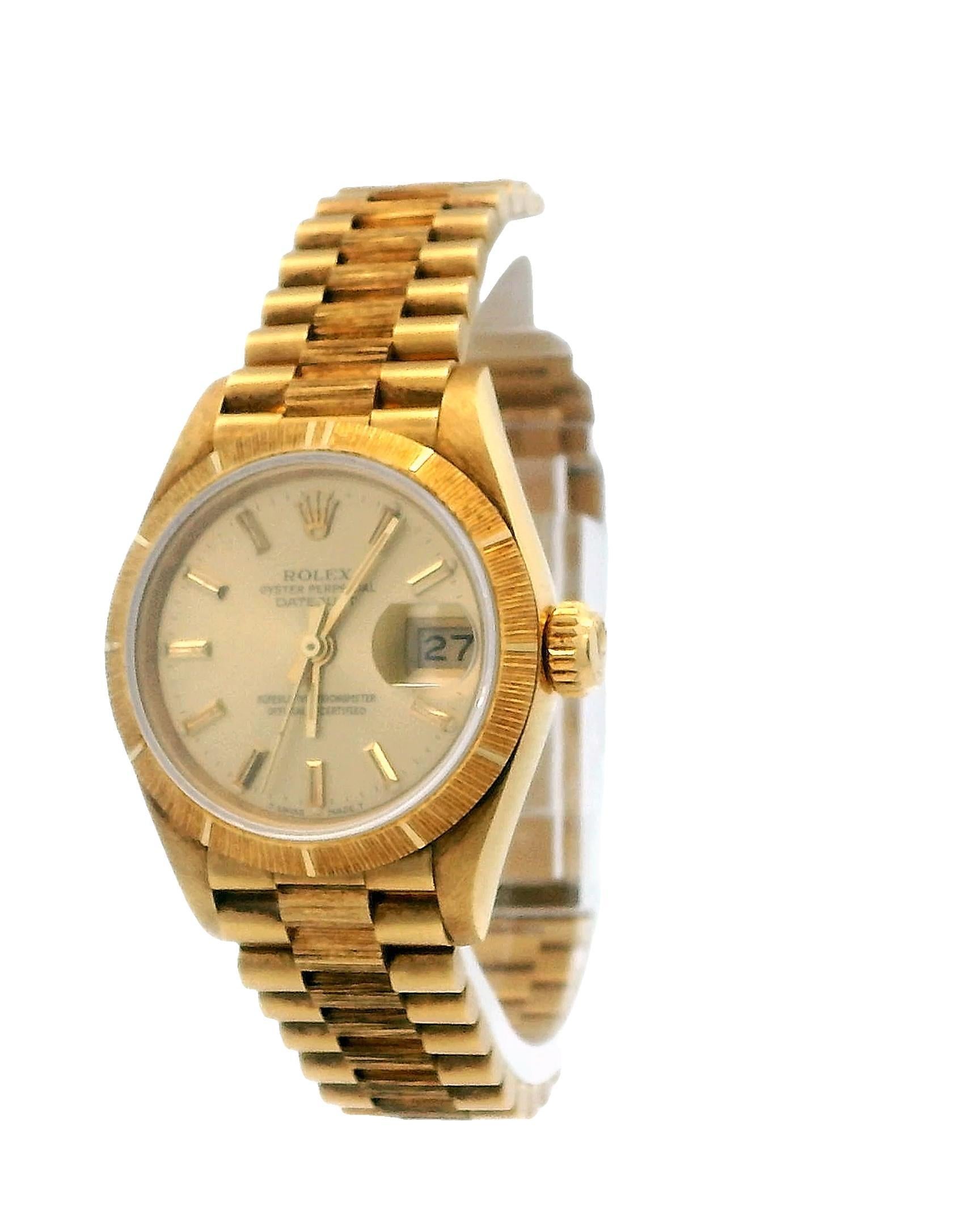 Rolex Datejust Ladies Watch 69278. This luxurious timepiece features a 26mm case made of 18k yellow gold, complemented by a stationary bezel with a bark finish. The champagne dial showcases gold hands and index hour markers, with the date displayed