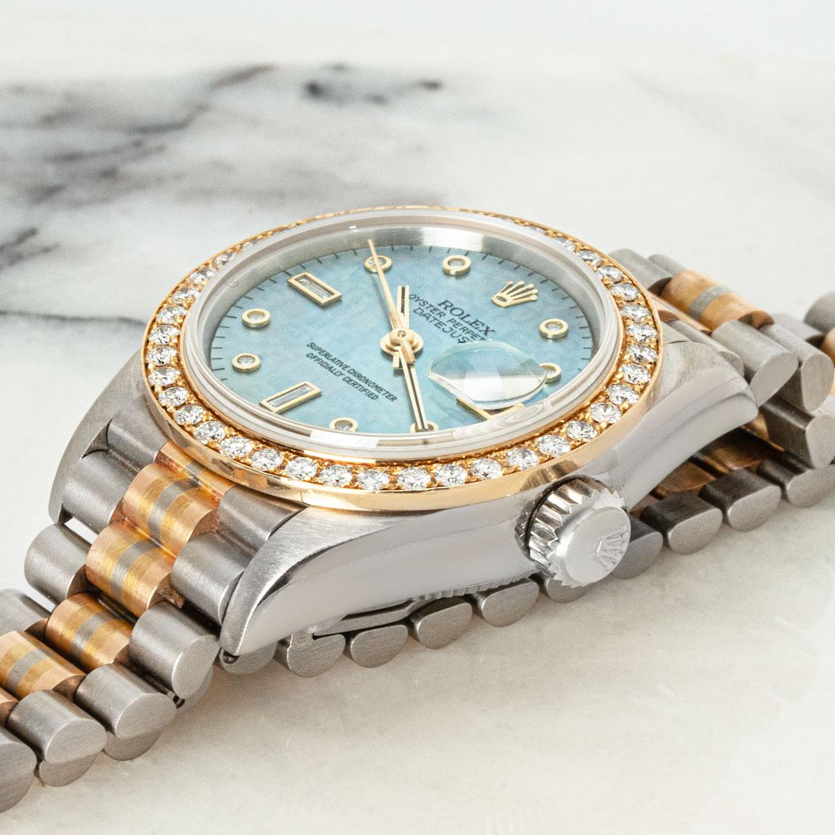 A 26mm Datejust in tri-gold by Rolex. Featuring a Tahitian mother of pearl dial with diamond set hour markers and a fixed yellow gold bezel set with 40 round brilliant cut diamonds.

Fitted with a sapphire crystal and a self-winding automatic