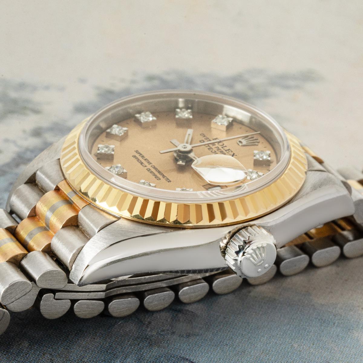 Eine 26-mm-Datejust aus Dreifachgold von Rolex. Das champagnerfarbene Zifferblatt mit diamantbesetzten Indexen wird durch eine feste, geriffelte Lünette aus Gelbgold ergänzt.

Der mit Saphirglas und einem Automatikwerk ausgestattete Zeitmesser wird