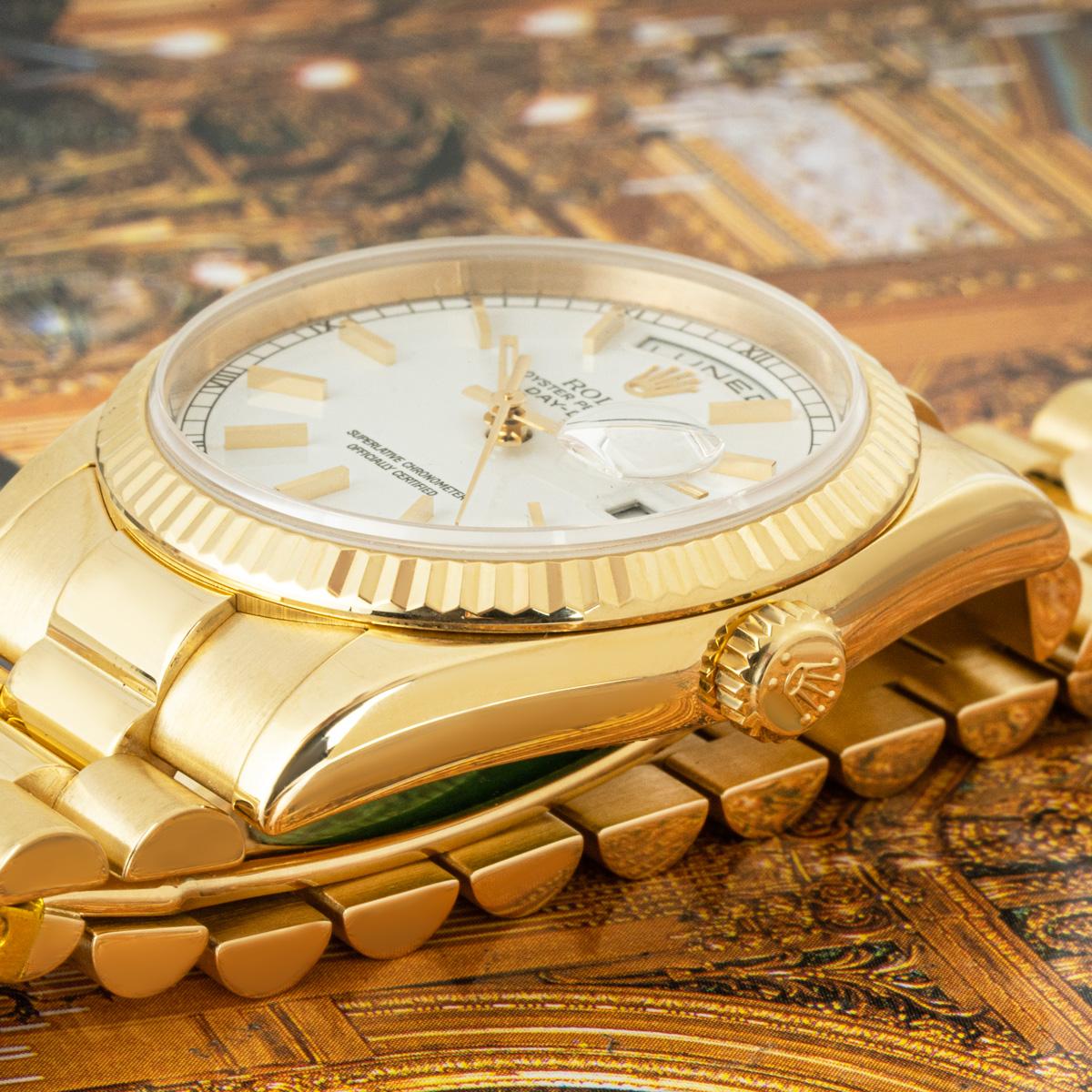 Eine Day-Date aus Gelbgold mit 36 mm Durchmesser von Rolex. Sie verfügt über ein weißes Zifferblatt mit applizierten Stundenmarkierungen sowie römischen Ziffern am äußeren Umfang des Zifferblatts und eine kannelierte Lünette aus Gelbgold. Das