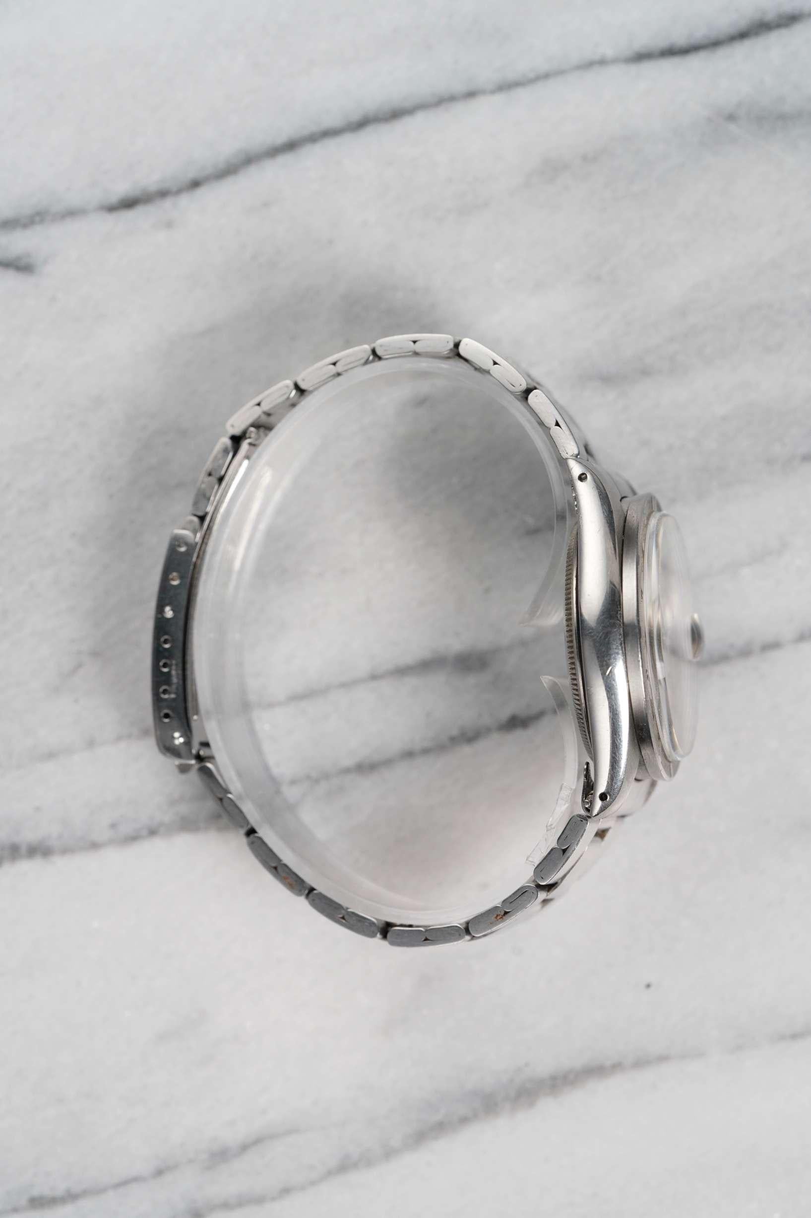 Voici une Rolex Oyster Perpetual référence 1501 automatique avec un cadran argenté à bandes verticales datant des années 1970. Boîtier en acier inoxydable de 34 mm avec lunette tournée et cornes percées. Monté sur son bracelet à rivets en acier, il