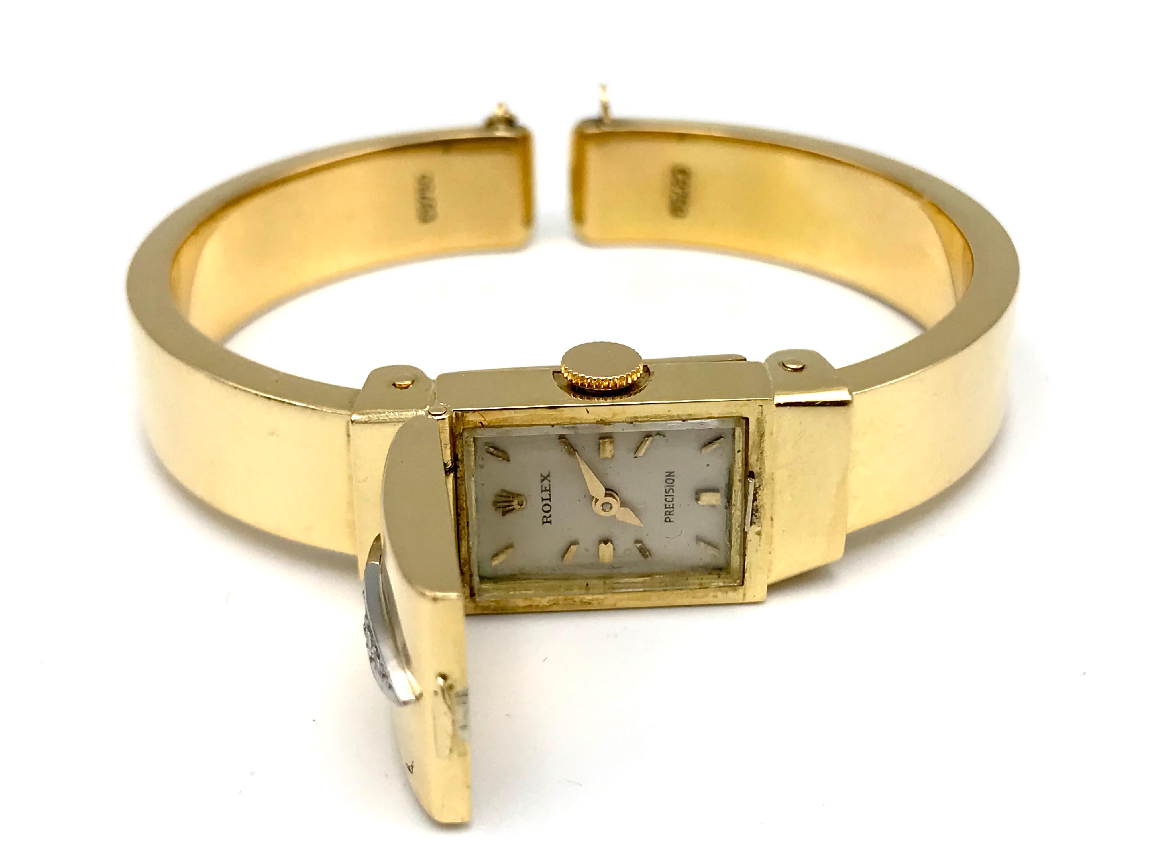 Stylish vintage (c. 1960s) wrist watch bracelet by Rolex. Made of 18k yellow gold, featuring diamond on top of the hinged cover. 
The watch is mechanical. Features gold hour and minute hands. The cuff bracelet has a safety chain. 
The dial signed