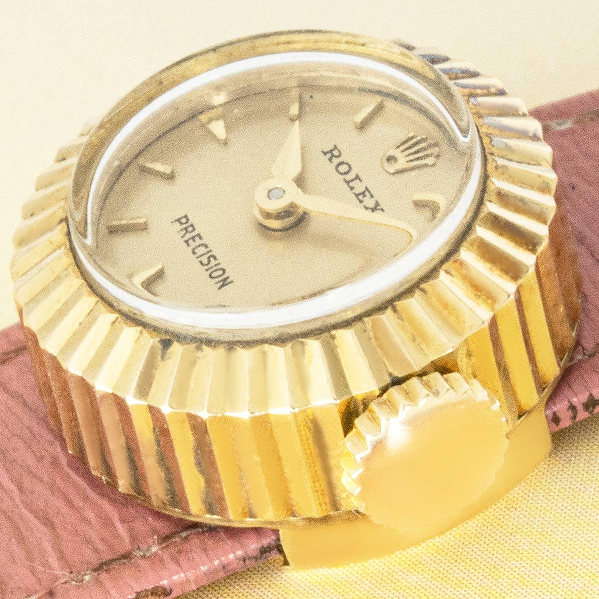 A vintage Precision Chameleon wristwatch in yellow gold by Rolex. Featuring a champagne dial with applied hour markers, complemented by a yellow gold fluted bezel.

Fitted with plastic glass and a manual wind movement. Presented on a pink Rolex