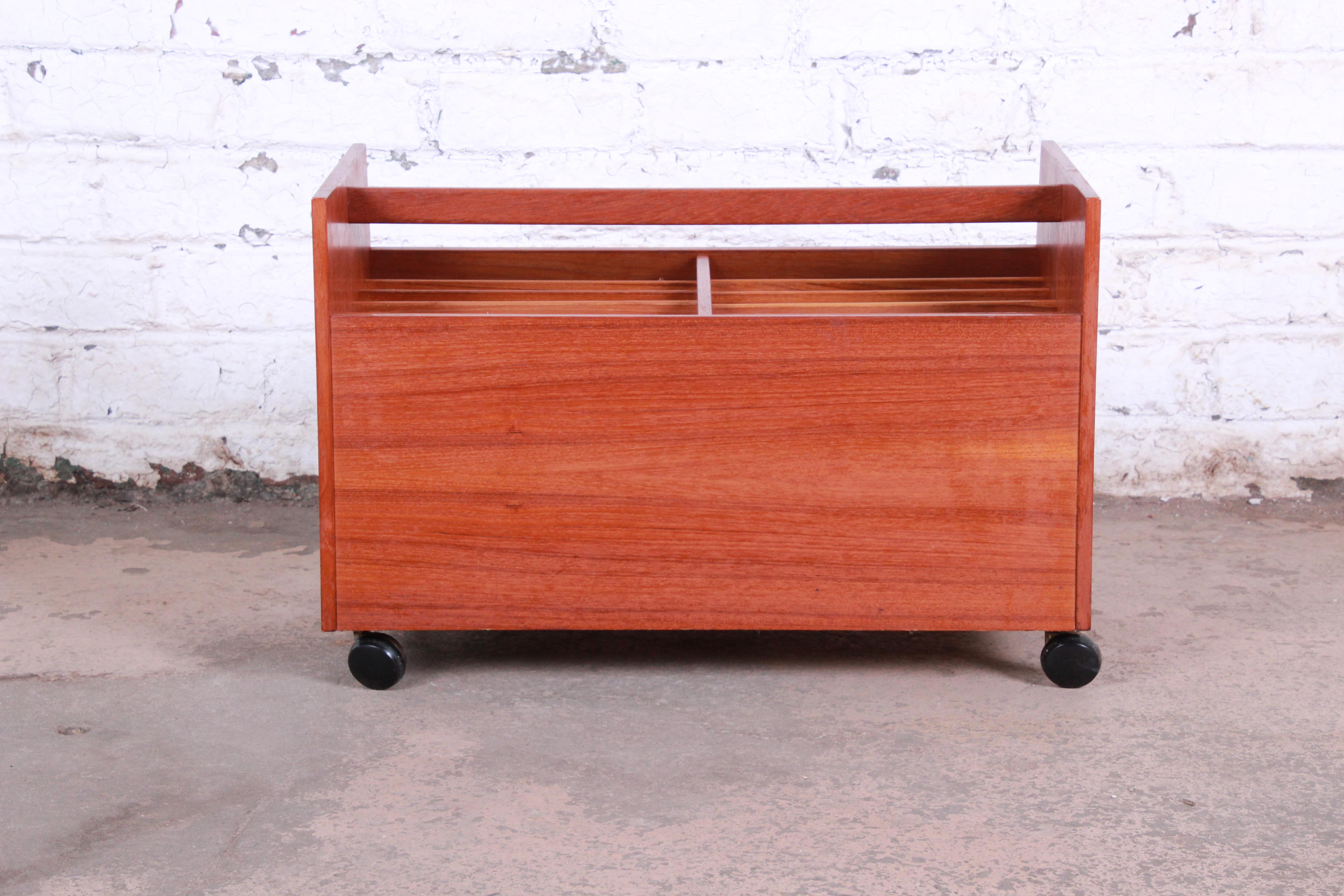 A gorgeous midcentury Norwegian modern record or magazine cart by Rolf Hesland for Bruskbø. The cart features stunning teak wood grain and sleek Scandinavian design. The trapezoidal shaped trolley has a handle on top and sits on rolling casters. The