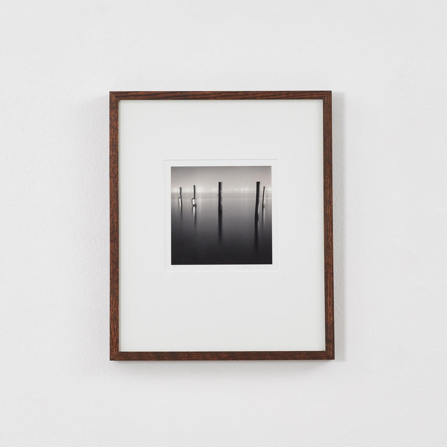 Cinq pilotis en bois marquent l'emplacement d'un dockland brumeux. Capturée par une longue exposition, la scène se dissout dans une quasi-abstraction, ses marques verticales rappelant peut-être les gravures sur bois japonaises éthérées de l'ukiyo-e.