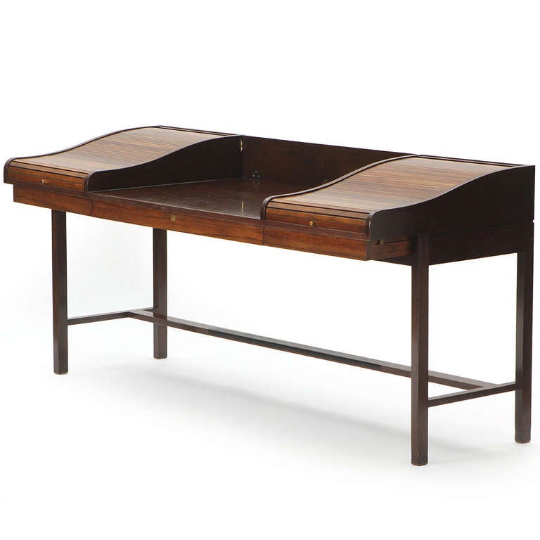 A stately and elegant rosewood and mahogany campaign desk having three drawers and two rosewood roll-top compartments flanking a central writing surface with a flip-down back splash, floating on a spare, architectural rectilinear base.