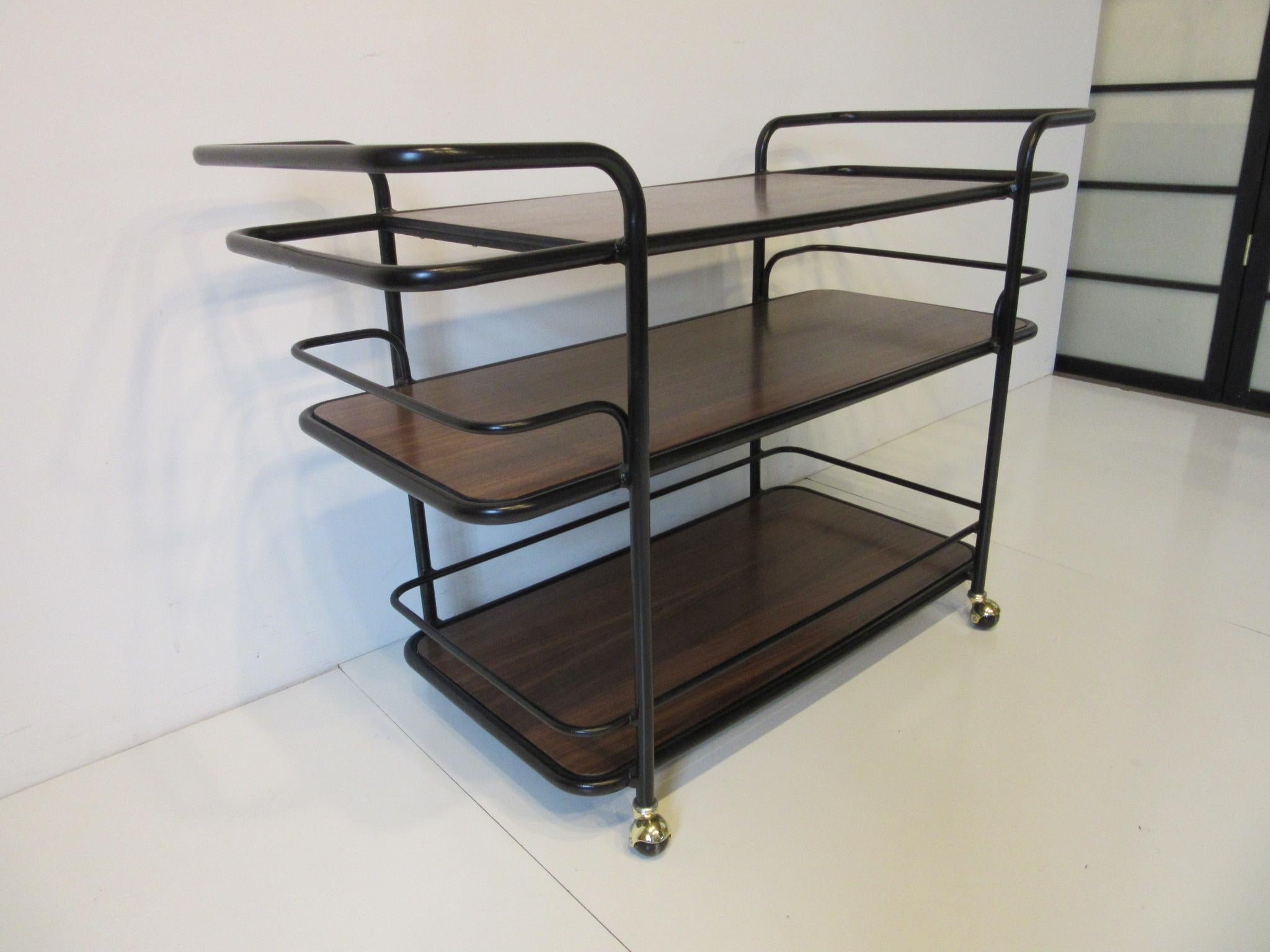 A satin black painted metal rolling bar cart with walnut shelves, brass wheels and each end having storage areas for bottles and glasses. In the manner of Art Deco and the International Style using the tubes and curves of that periods design