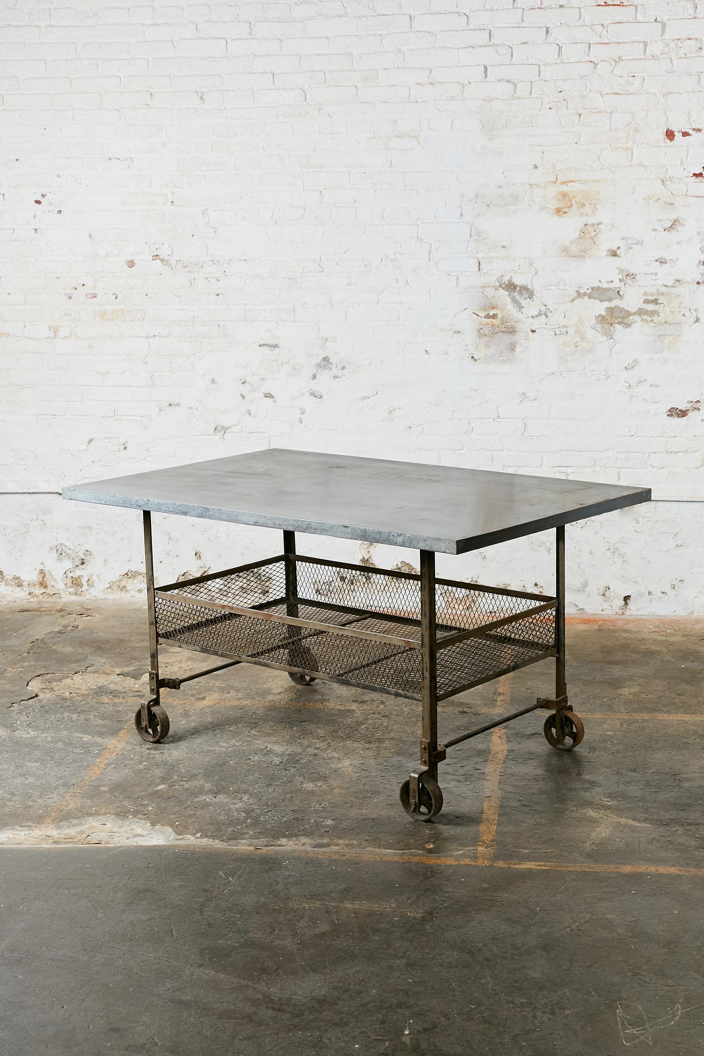 Rolling industrial table. Top covered in zinc and shelf/basket made of wire. All four legs rest on pivot wheels.