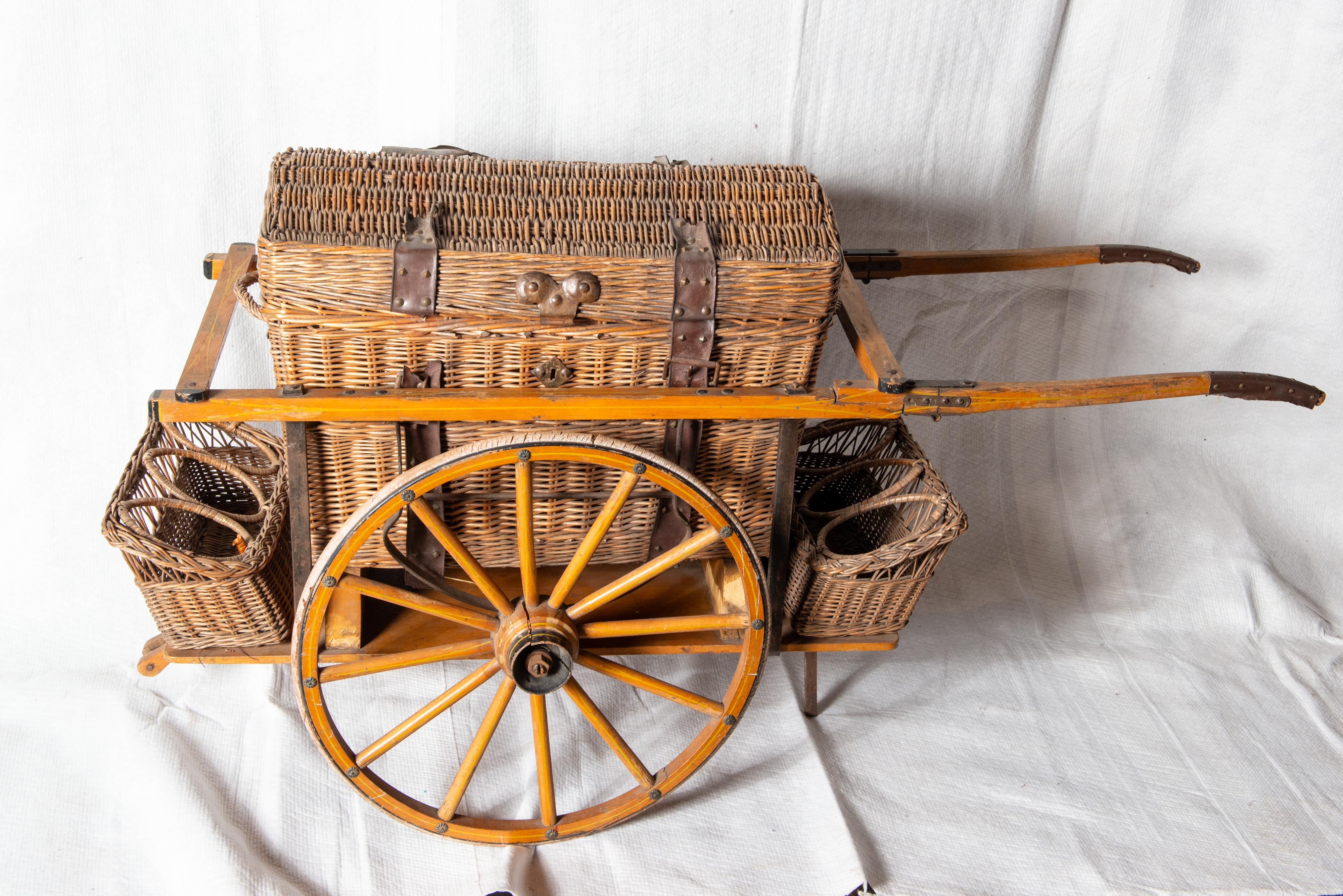 A very unusual antique wicker picnic basket set in a large wheeled cart. The cart has six circular sections for wine or water bottles. The deep covered wicker basket has a handle on each end. Inside the basket there is a wood tray with a wicker