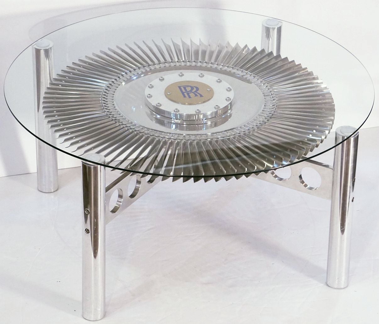 A stylish low cocktail or coffee table from England, featuring a round glass top (41 3/4 inches diameter) set upon a base with an authentic Rolls Royce rotating impeller from a British military jet engine, mounted to a four-post frame of tubular