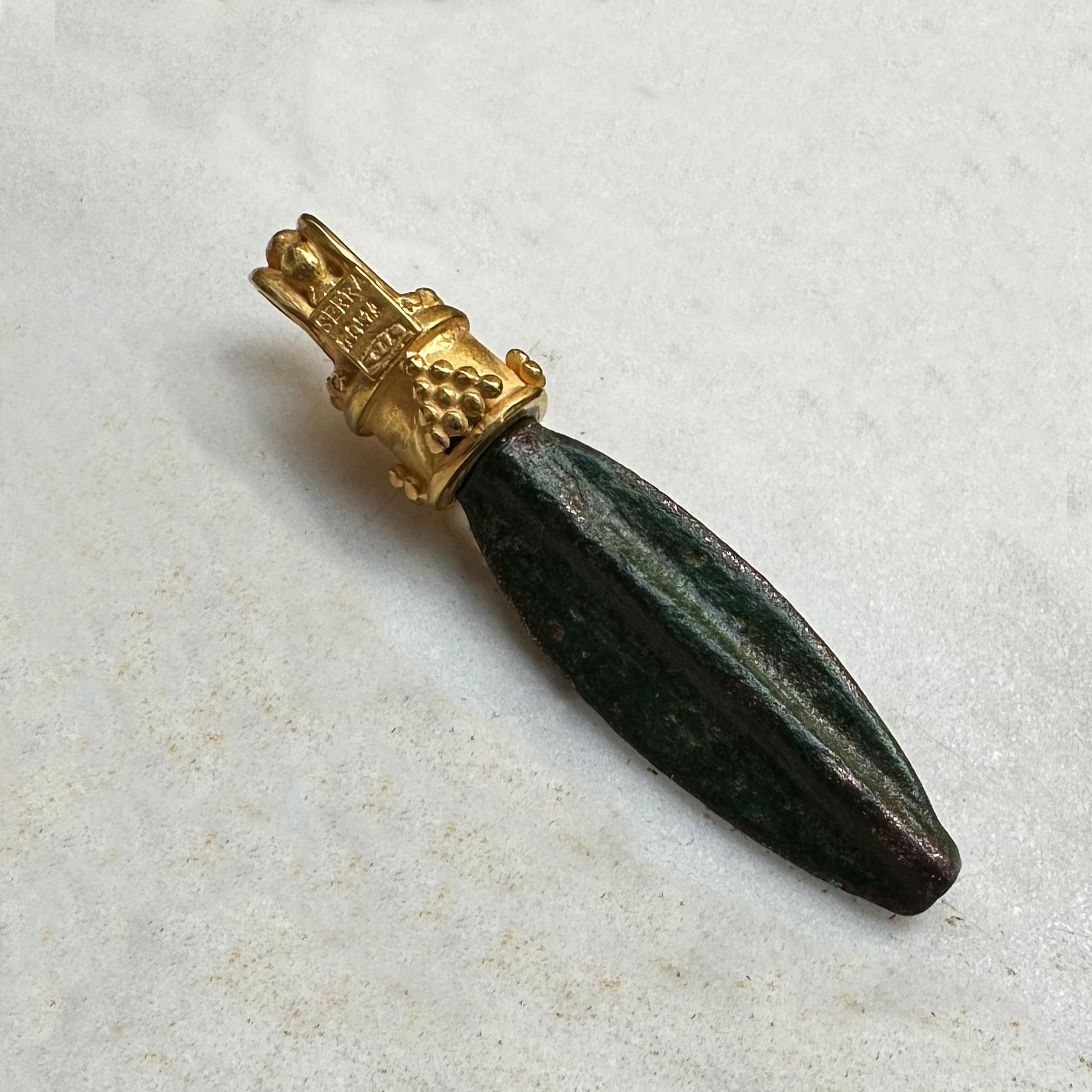 This small bronze arrowhead, exhibiting an olive-green patina, resembles examples from the Villa Julia Collection. It is mounted in 18-karat gold, showcasing an exclusive design by the Serra store. The most common form of Roman arrowhead was