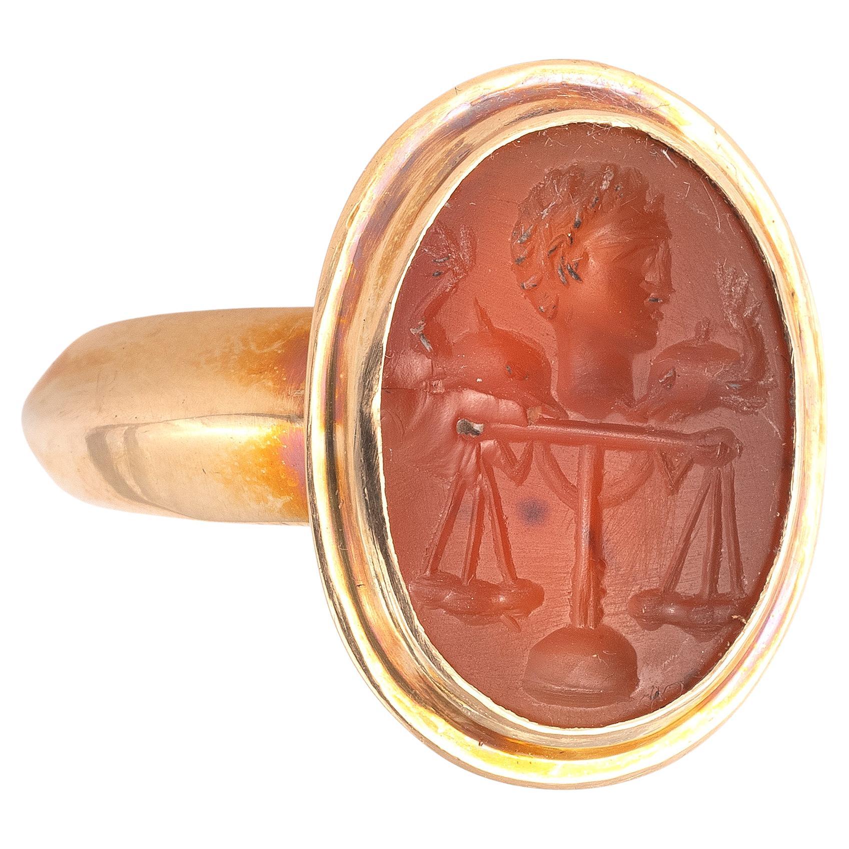 Oval intaglio engraved with a balance surmounted by the profile of Arione, son of Poseidon, flanked by two dolphins. 
The myth presents Arione as an important cither musician, saved from the dolphins in the open sea. The orange carnelian intaglio