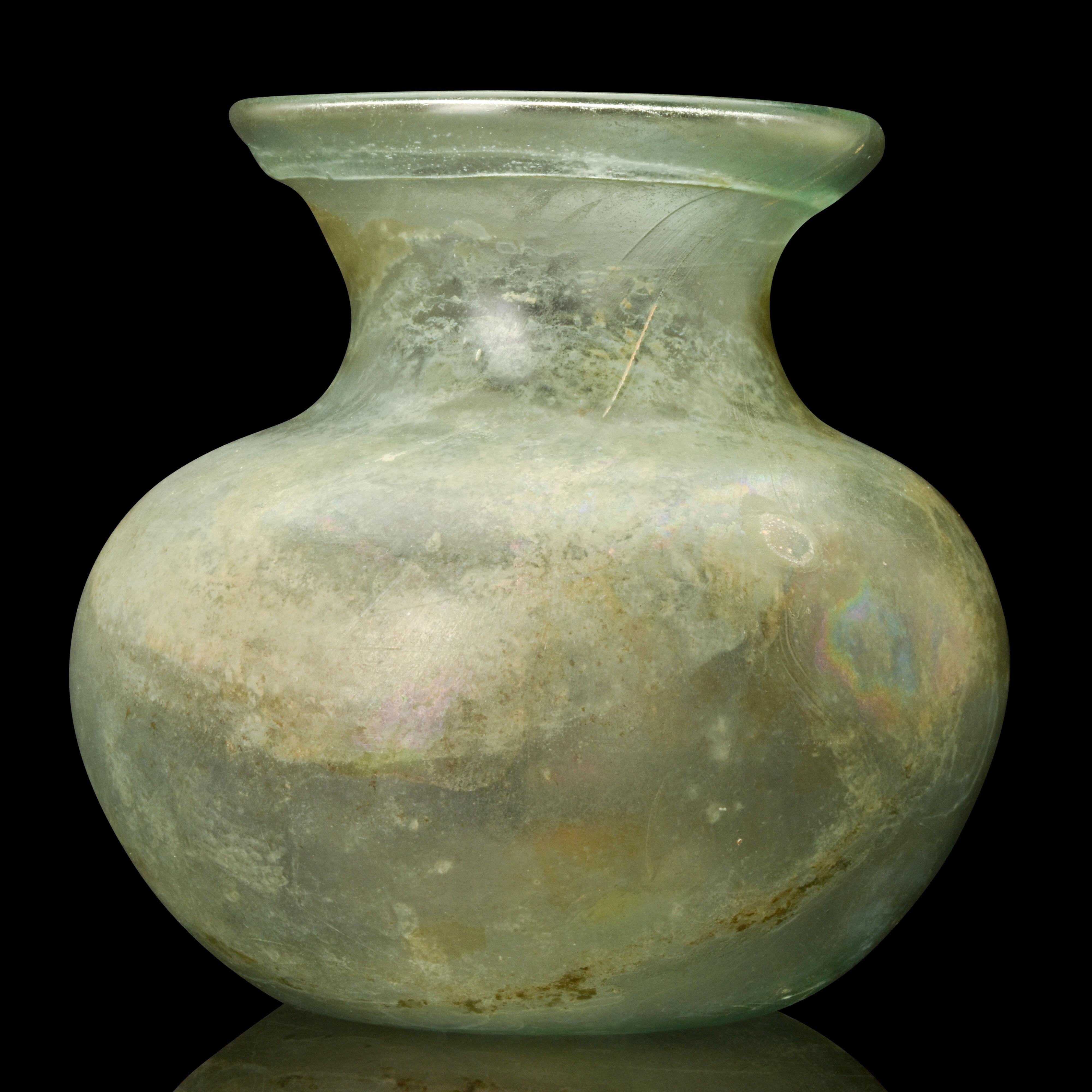 A beautiful Roman translucent glass jar, with a light blue-green tinge. It has a thick, splayed rim that is folded over and into a short, concave neck. The body is squat and bulbous, with a slightly pushed-in bottom. Throughout the vessel, there are