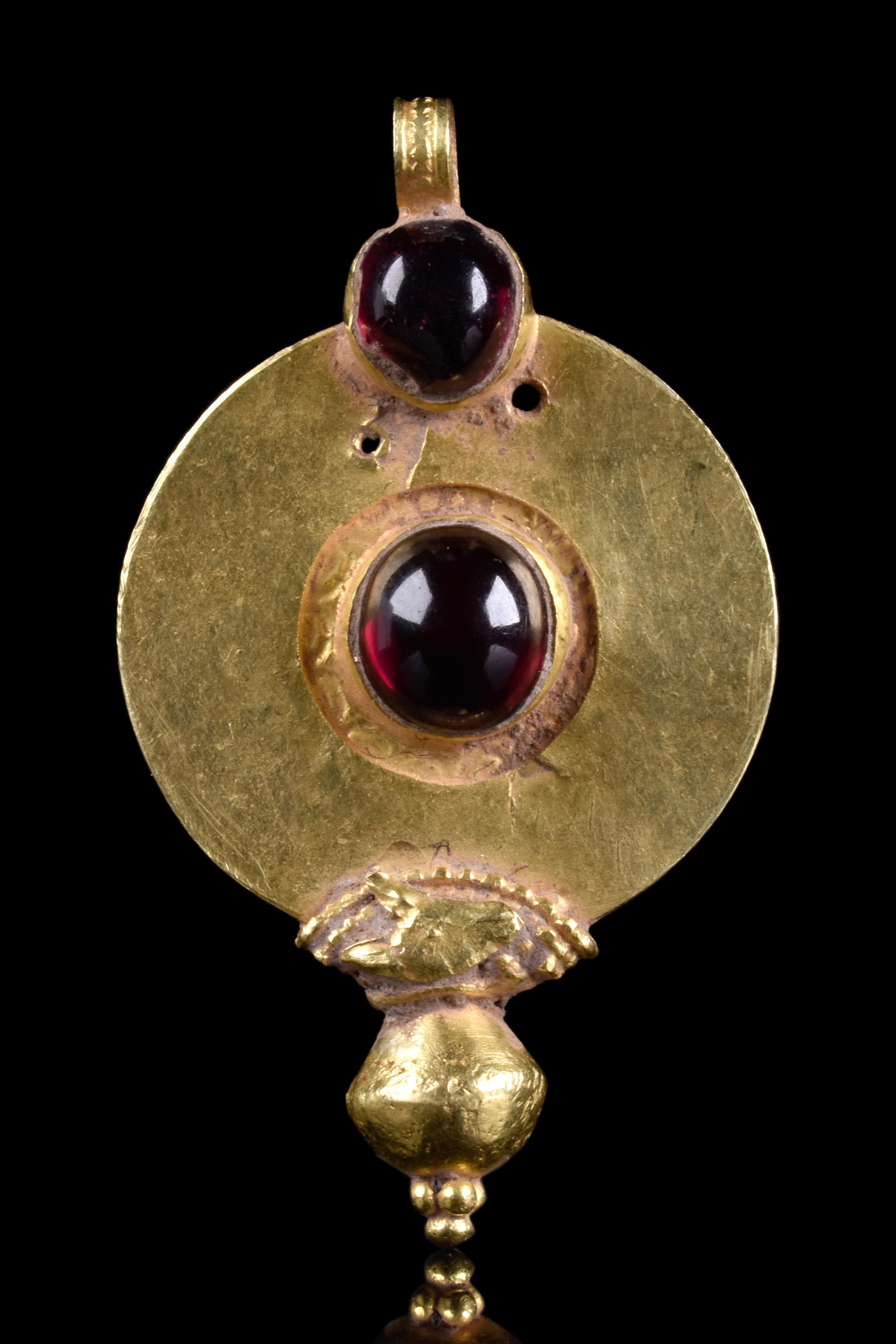 A thin, circular Roman gold pendant featuring a central garnet cabochon gem held within a raised cell. Above it, another garnet cabochon enriches the piece. The item is topped by a suspension loop. At the bottom, a biconical decoration with filigree