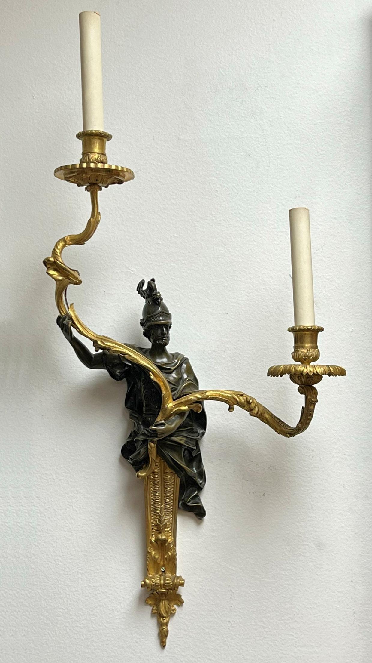 Our pair of gilt bronze sconces with the patinated figures of Greek or Roman warriors Measure 23 by 13 by 6.75 inches and have two candle arms with candelabra Size sockets. Apparently unsigned. In need of new wiring.