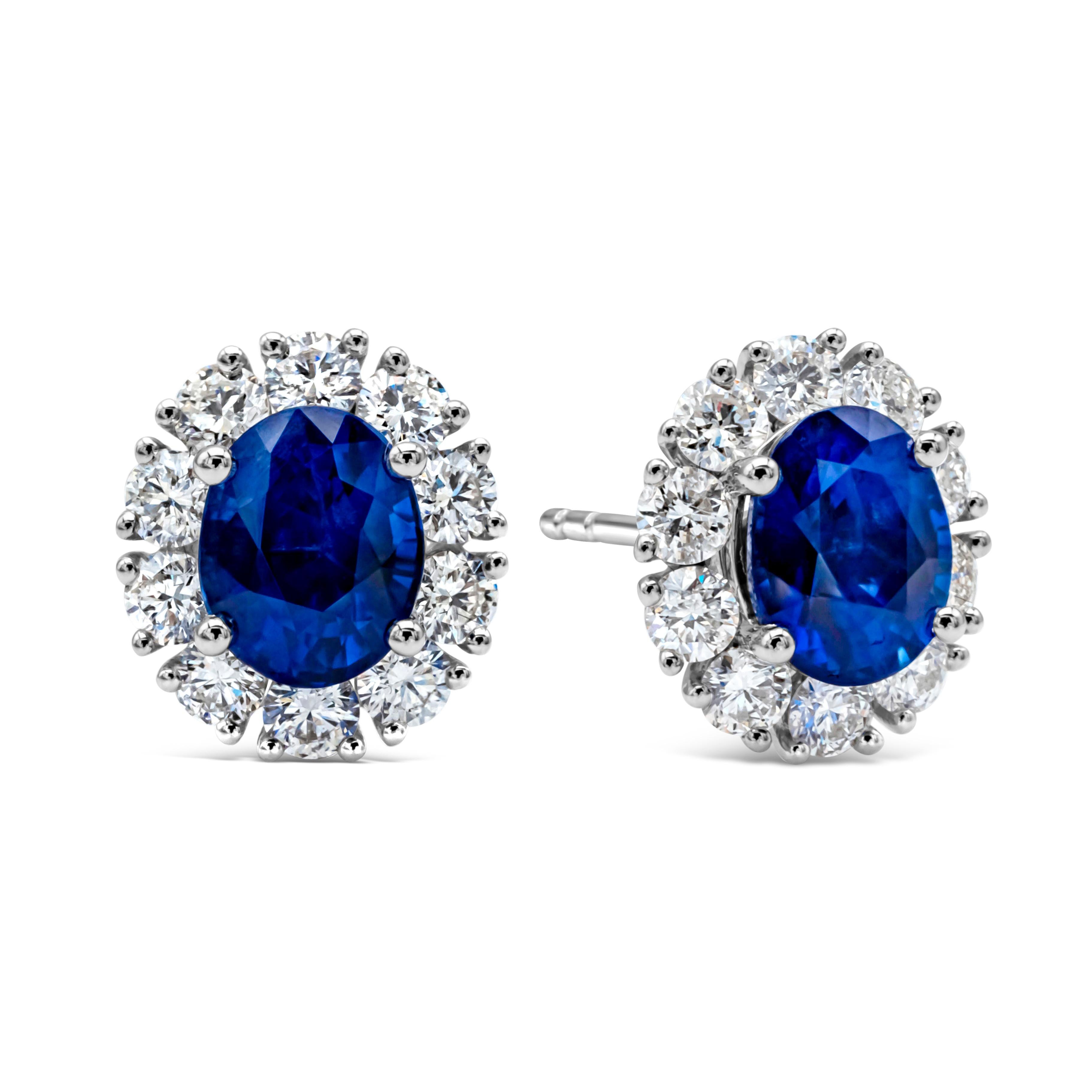 A simple and classic pair of stud earrings showcasing an oval cut blue sapphires weighing 2.29 carats total, set in a timeless four prong basket setting. Surrounded by a single row of round brilliant diamonds weighing 0.84 carats total, F color and